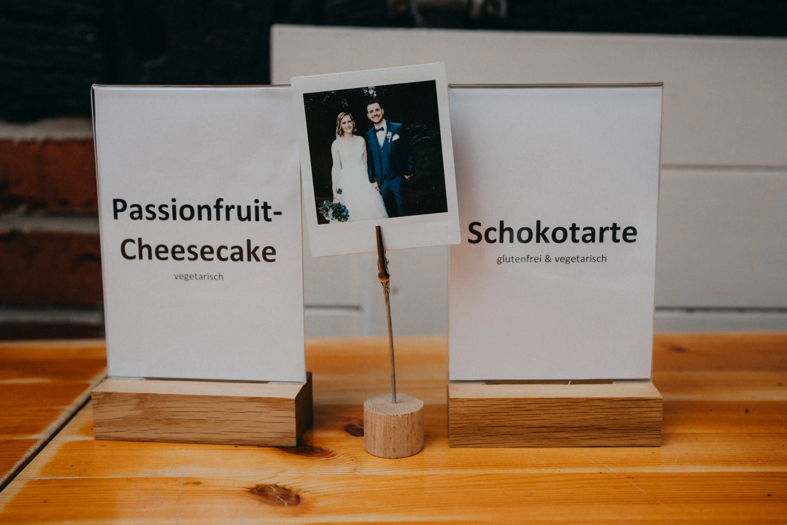 Zwei weiße Schilder auf Holzsockeln, eines mit dem Schriftzug "Passionfruit-Cheesecake" und das andere mit "Schokotarte", beide in einer Vitrine. Zwischen den Schildern liegt eine Fotokarte eines lachenden Paares in formeller Kleidung. Das Ganze steh