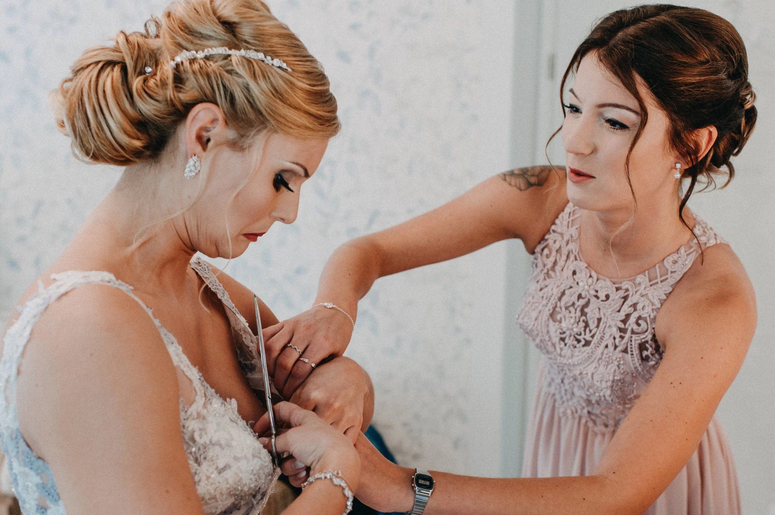 Eine Braut in einem weißen Spitzenkleid wird von einer anderen Frau angeleitet, während sie eine Halskette anlegt. Die Frauen tragen elegante Kleider und haben sorgsam gestylte Frisuren und Schmuck. Die Stimmung ist andächtig und feierlich.