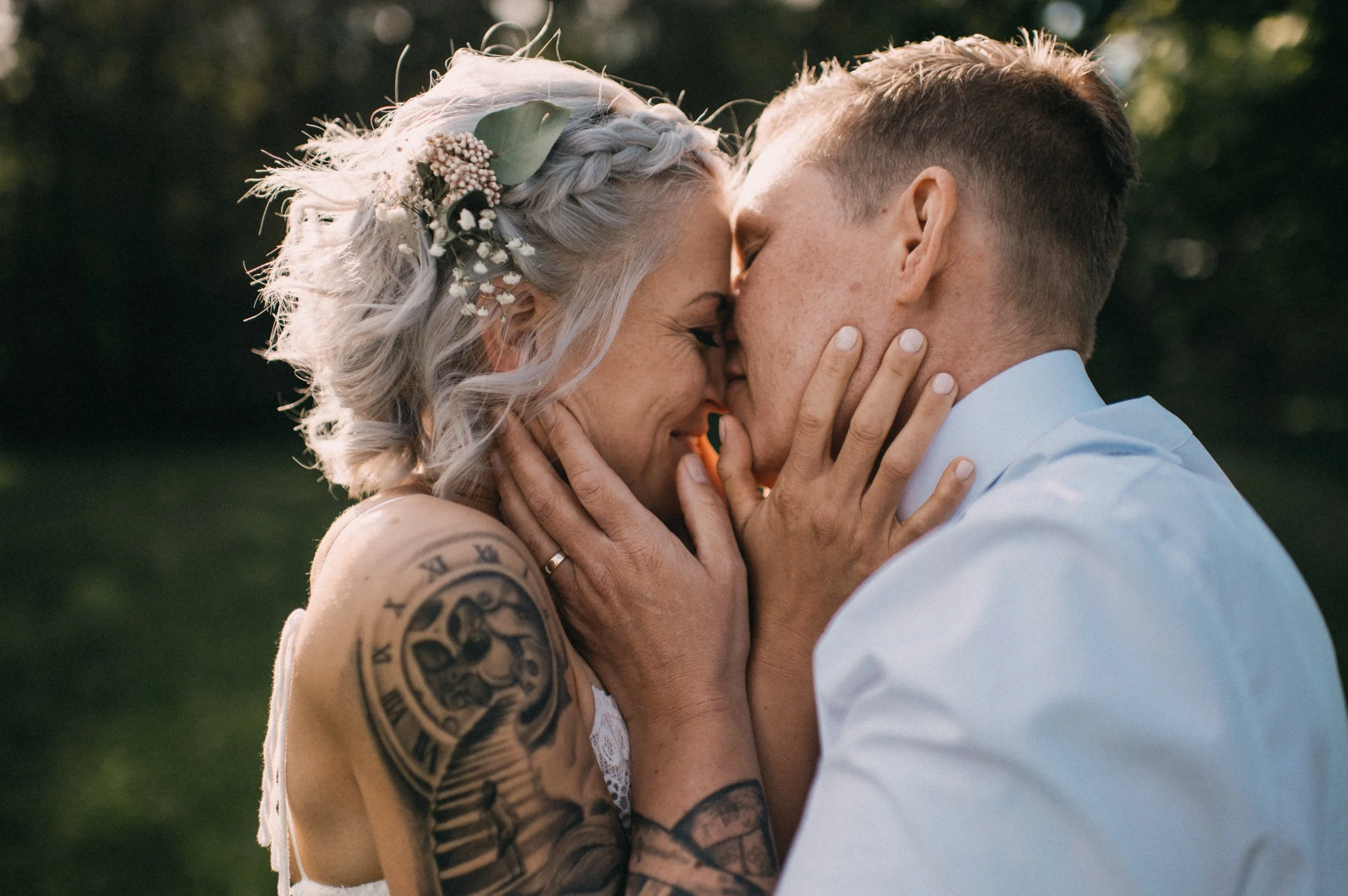 Ein Paar beim Kuss, im Freien, bei Sonnenuntergang, die Frau trägt einen Haarschmuck mit Blumen und Blättern, hat Tattoos am Arm, der Mann trägt ein weißes Hemd.