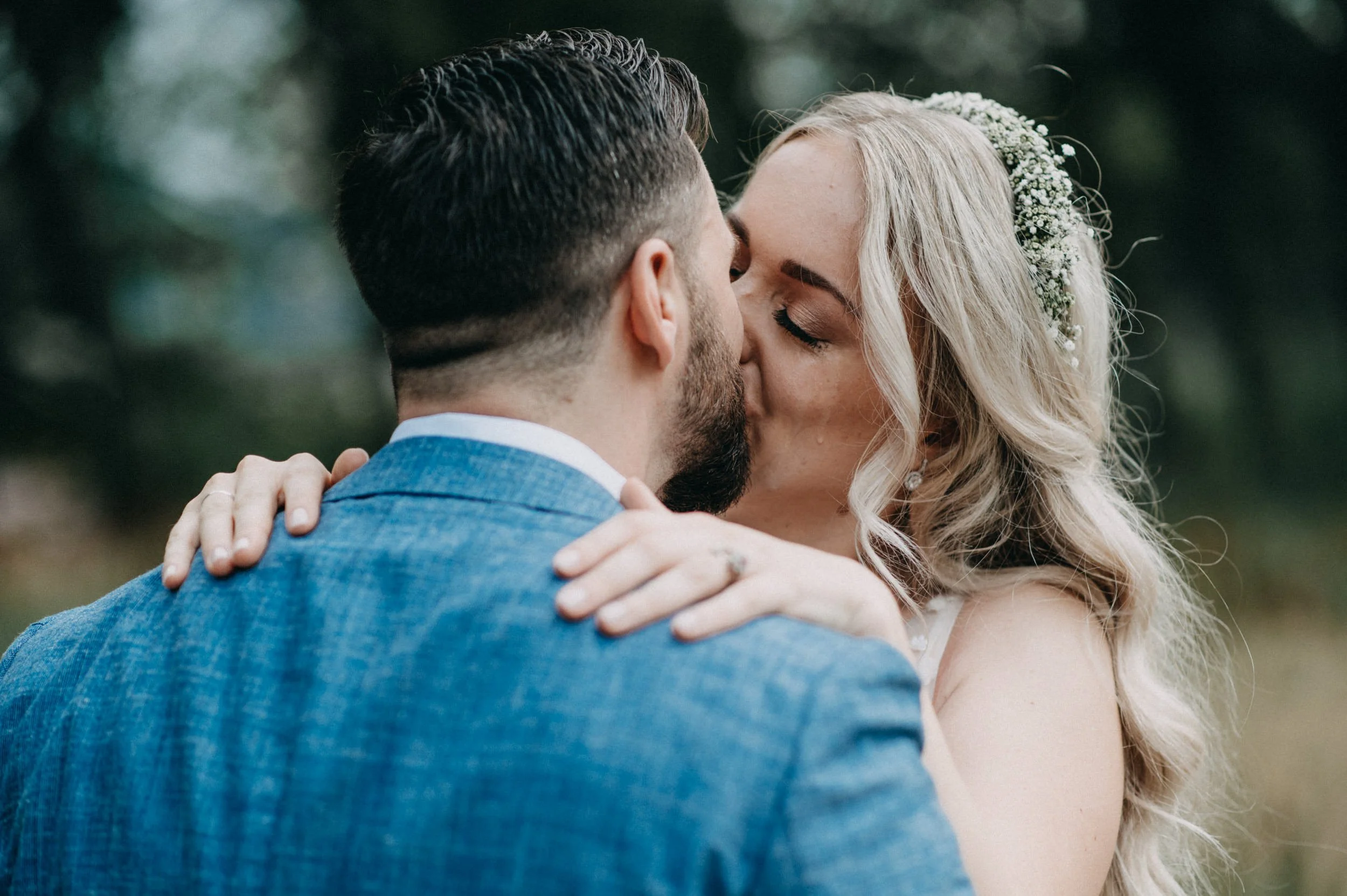 Ein Herzstück einer Hochzeitszeremonie, bei der ein Mann und eine Frau sich küssen, der Mann trägt einen blauen Anzug, die Frau trägt einen weißen Kleid mit Blumenschmuck im Haar, im Hintergrund ist eine grüne Naturszene.