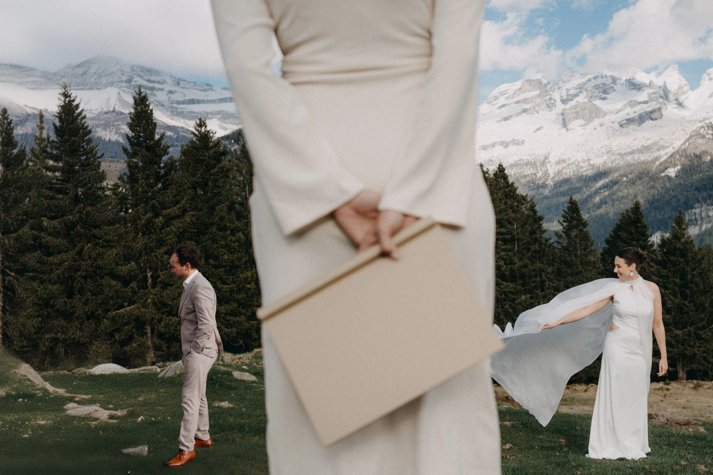 Ein Brautpaar im Freien bei einer Hochzeit, im Hintergrund schneebedeckte Berge und Tannenbäume, zwei Personen, einer männlich in Anzug, einer weiblich in weißem Brautkleid, tanzen.