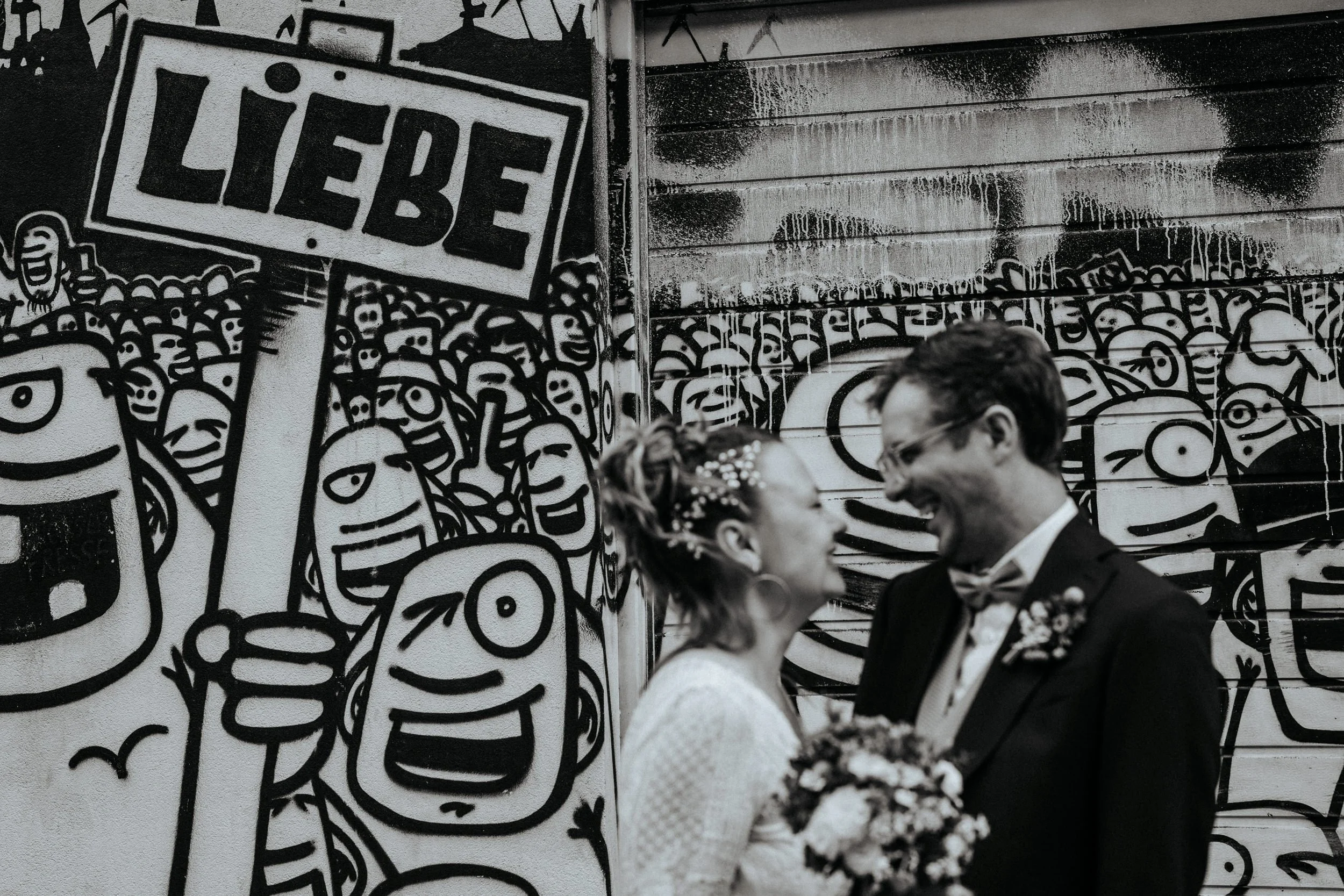 Ein Hochzeitsbild von einem Brautpaar, das sich glücklich ansieht, vor einer Wand mit Graffiti-Kunst.