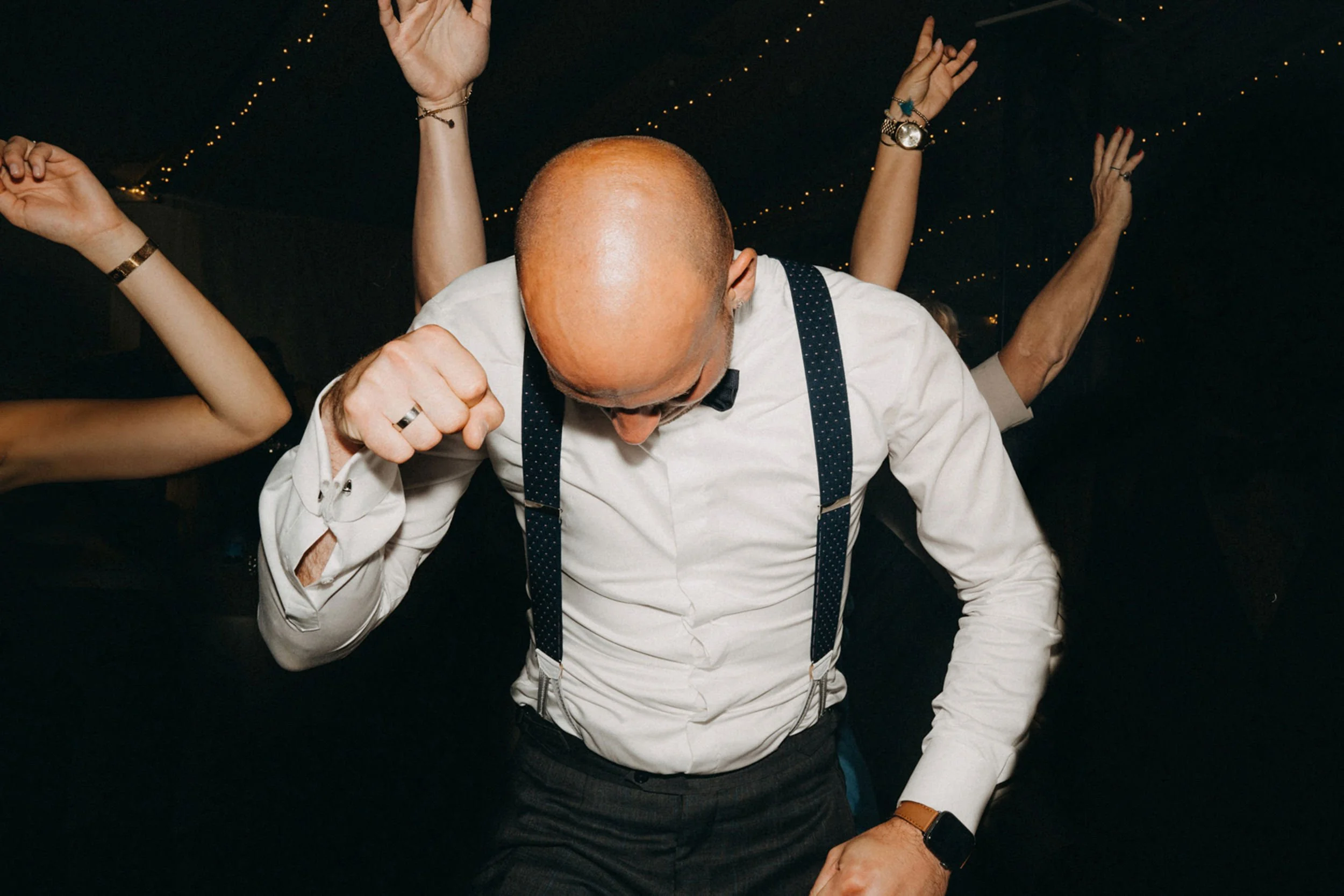 Person in formeller Kleidung tanzt mit anderen auf einer Party bei Nacht, im Hintergrund Lichter. Der Blick ist auf den Kopf und die Brust gerichtet.