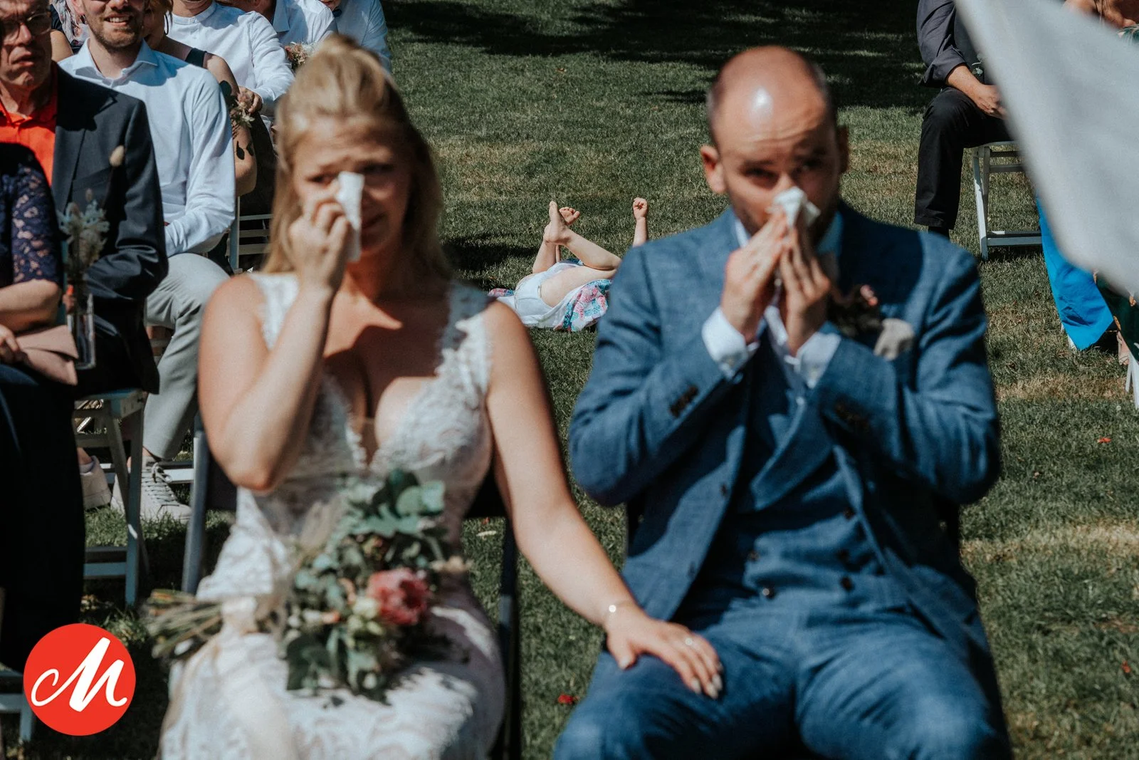 Ein Brautpaar bei einer Hochzeit sitzt im Freien, beide weinen und wischen sich die Tränen mit Taschentüchern.