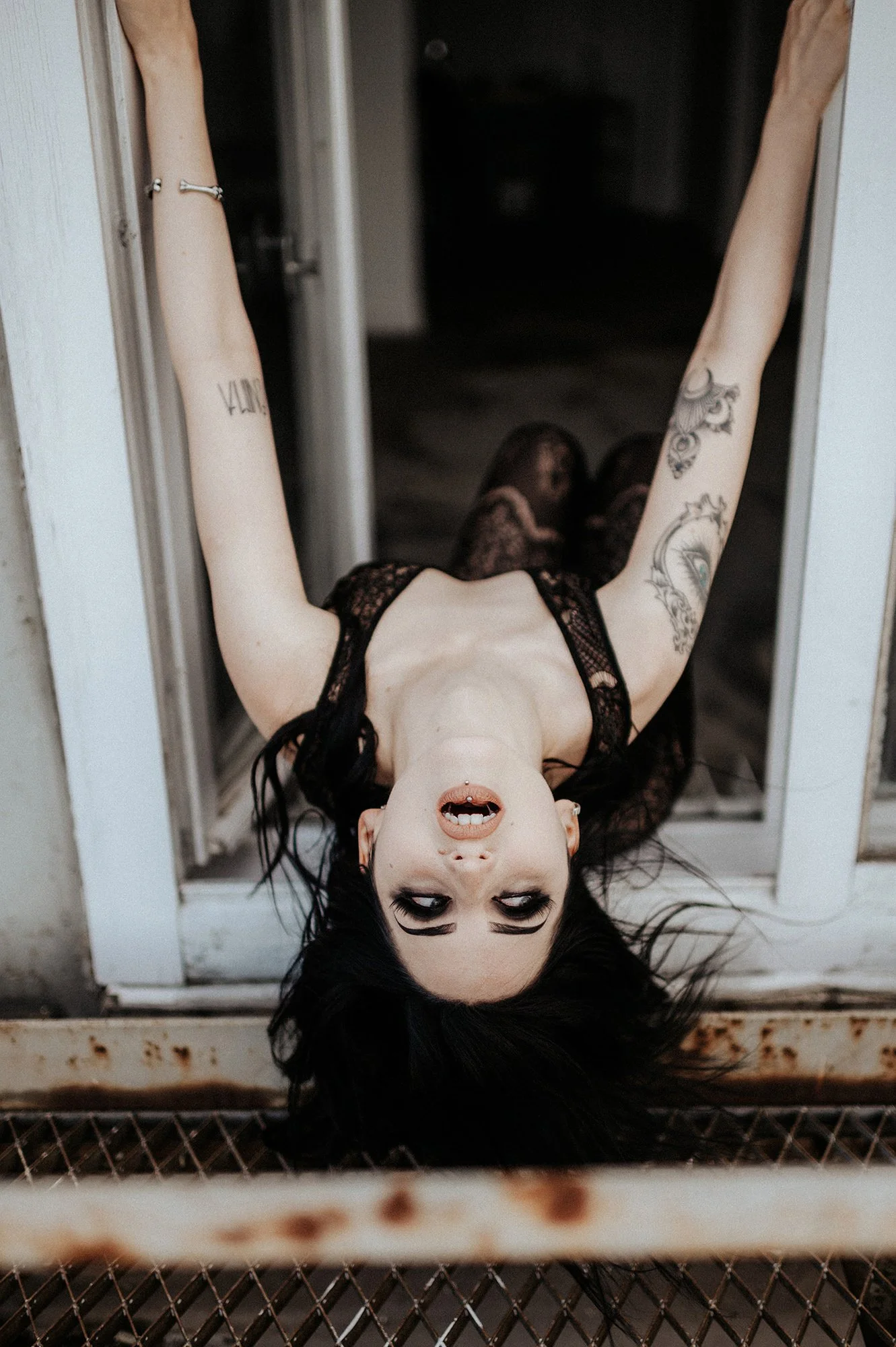 Eine Frau liegt mit dem Kopf nach unten auf einem Geländer, Blick nach oben, trägt schwarze Haare, dunkles Make-up und einen dunklen Kleidungsstil, Tattoos an den Armen, in einer Umgebung mit rauem, industriellem Ambiente.