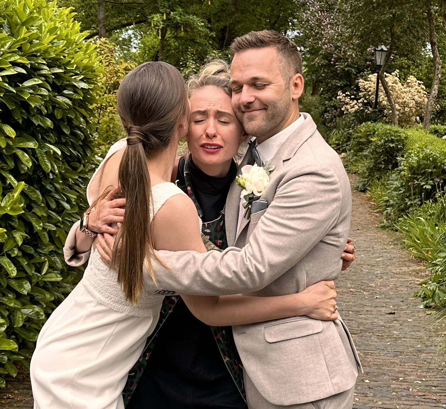 Drei Menschen umarmen sich herzlich im Garten, eine trägt ein weißes Kleid, der Mann trägt einen Anzug mit einer Blume im Revers, die Frau in der Mitte scheint emotional berührt zu sein.