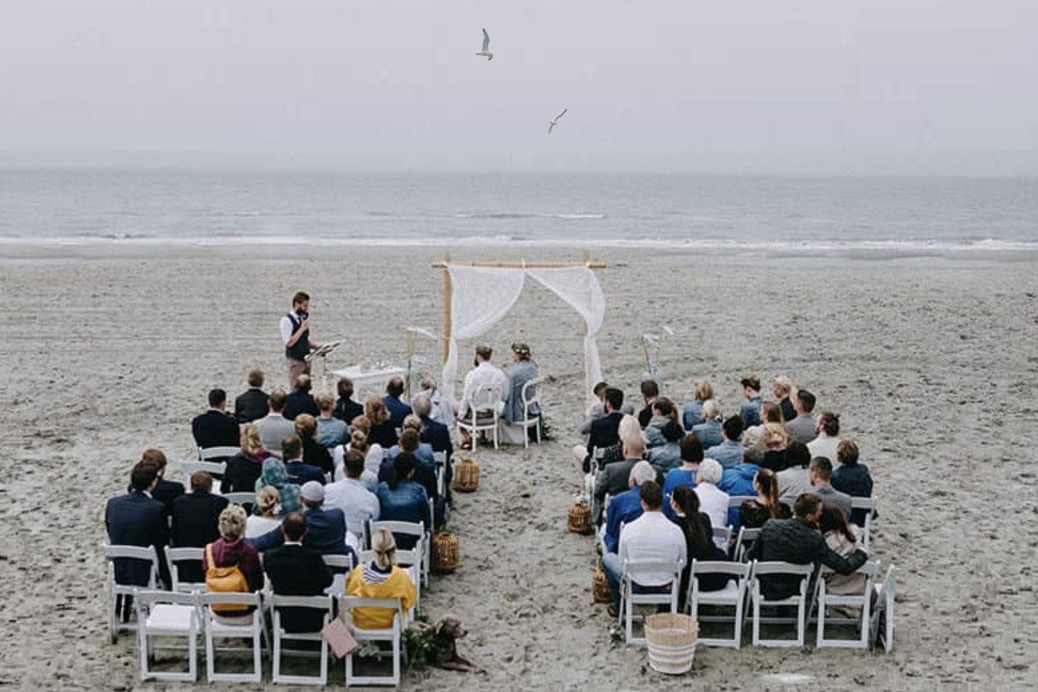 Eine Strandhochzeit mit Gästen, die vor dem Meer sitzen, während das Paar unter einem weißen Holz- und Stoffkabinett heiratet.