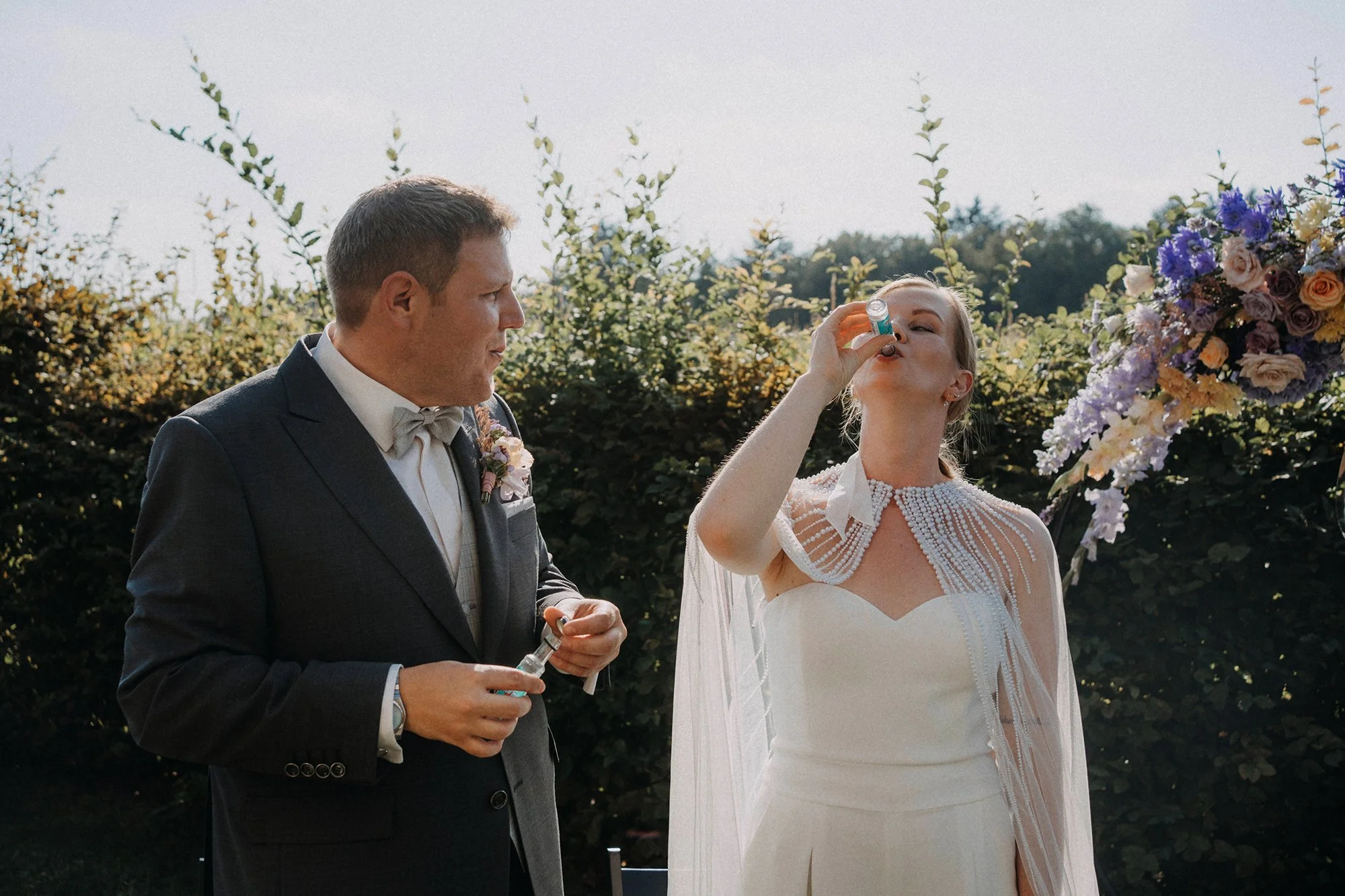 Ein Mann in einem schwarzen Anzug mit Fliege steht neben einer Frau in einem weißen Brautkleid bei einer Hochzeit im Freien. Die Frau trinkt aus einem kleinen Fläschchen, während sie neben einem großen Blumenarrangement steht.