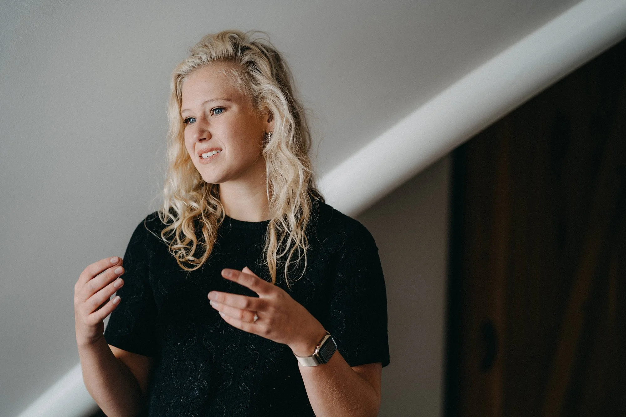 Eine junge Frau mit blonden, lockigen Haaren, trägt einen schwarzen Pullover und einen Smartwatch. Sie steht im Raum und scheint zu sprechen oder zu erklären.