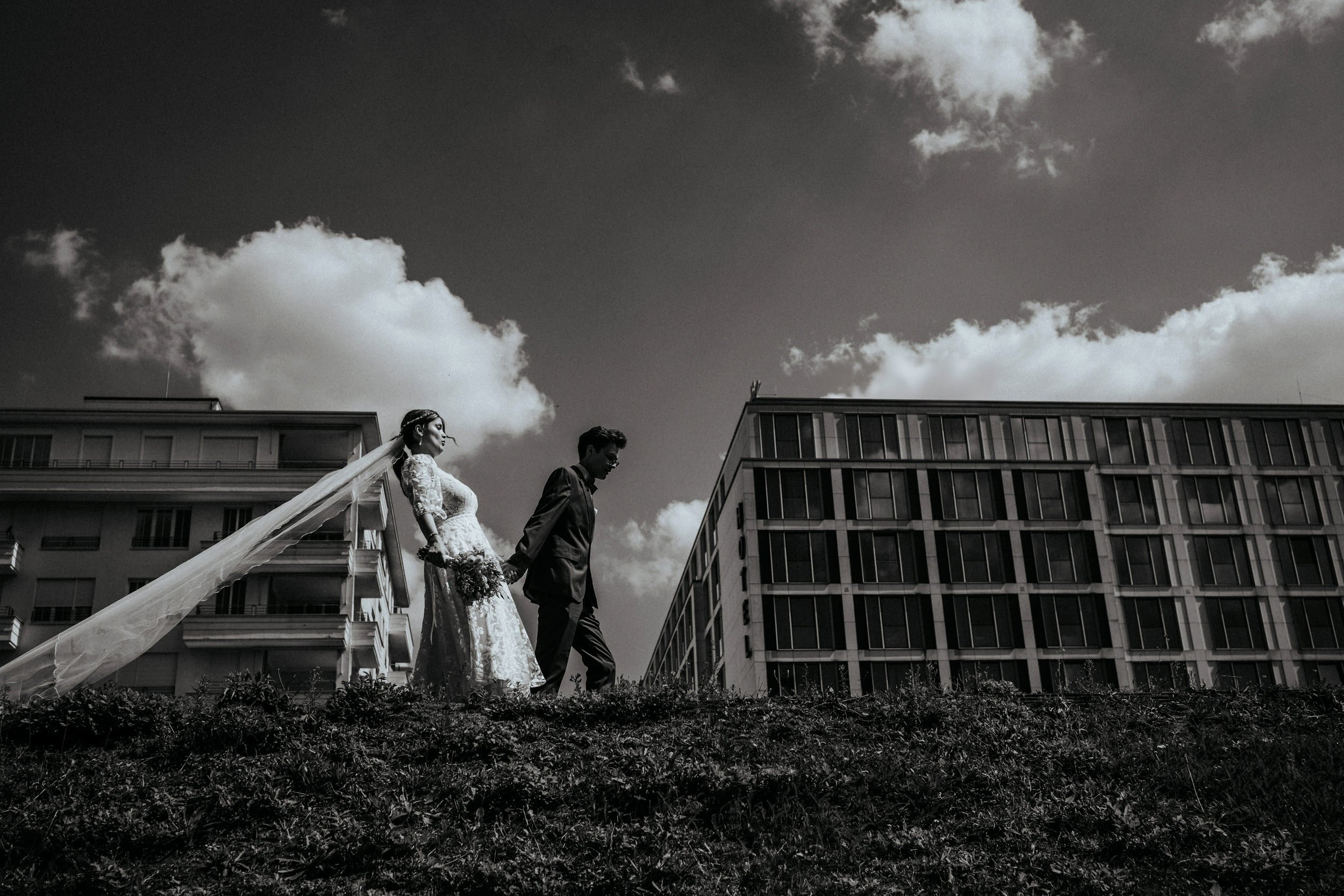 Schwarz-weiß-Fotografie eines Paares bei einer Hochzeit, das auf einer Wiese vor modernen Gebäuden steht, Himmel mit Wolken im Hintergrund.