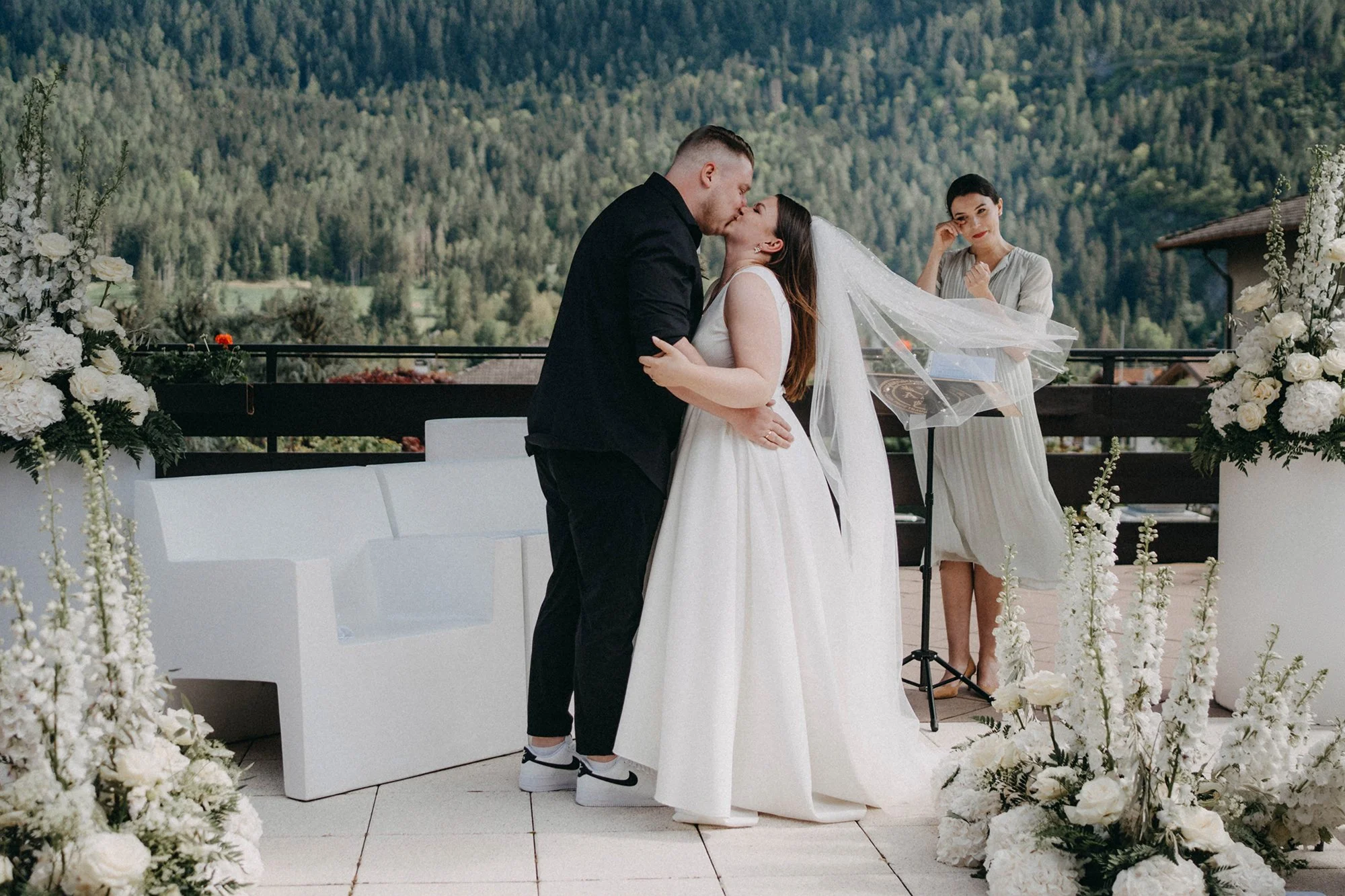 Ein Brautpaar küsst sich während ihrer Hochzeit auf einer Terrasse mit Bergblick, umgeben von weißen Blumen. Eine Frau steht neben ihnen, beobachtet die Szene.