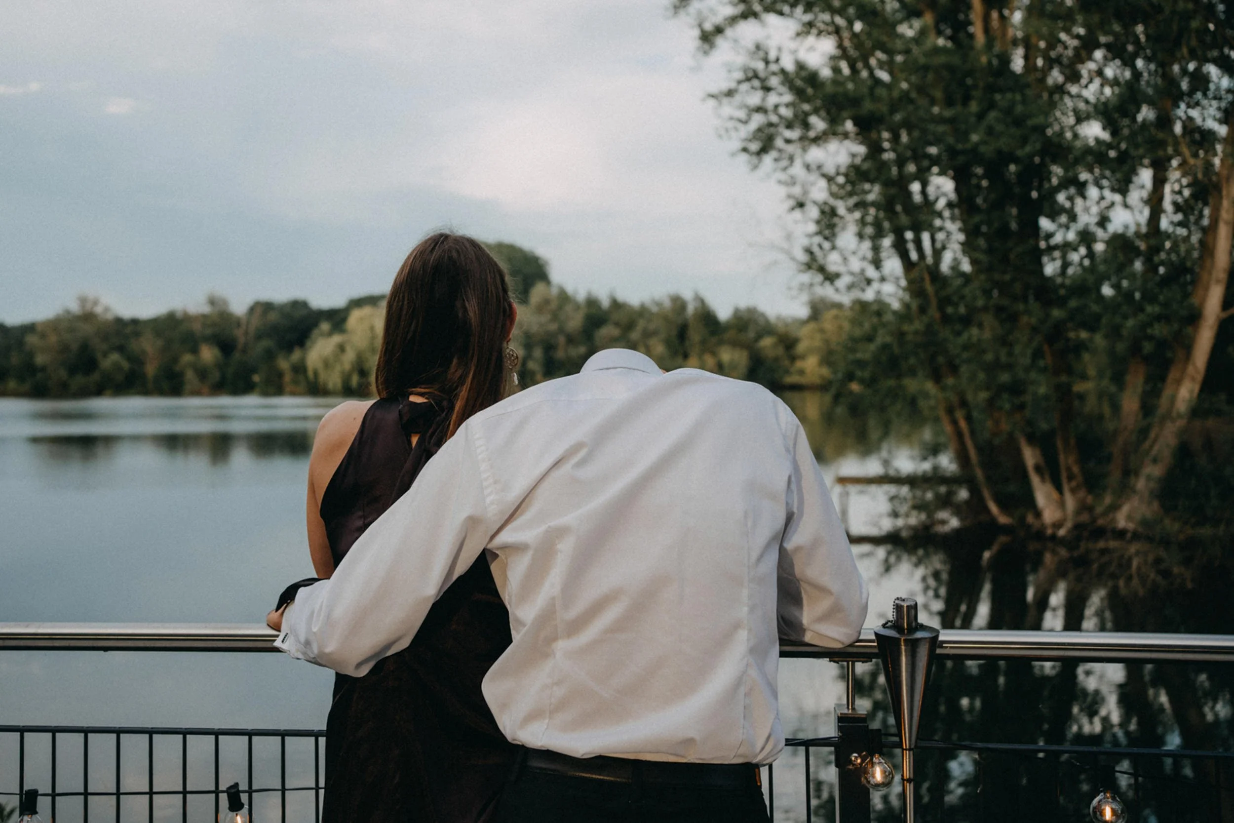 Zwei Menschen sitzen an einem Geländer am Wasser, einer trägt ein weißes Hemd, die andere eine schwarze Kleidung. Es ist dusk in einer ruhigen, naturreichen Umgebung mit Wasser und Bäumen.