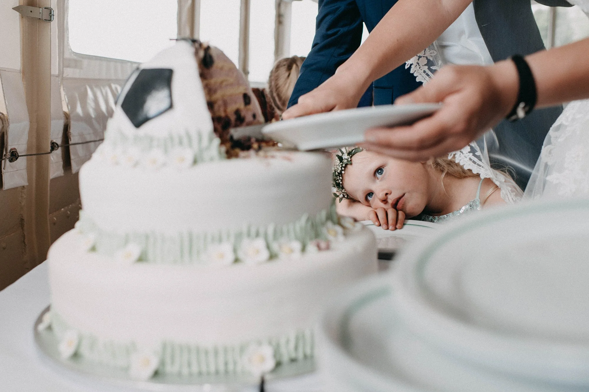 Ein Mädchen mit blauen Augen und Kranz aus Blumen im Haar liegt auf einem Tisch und schaut auf eine große, mehrstöckige Torte mit weißen Blüten und grünem Dekor.