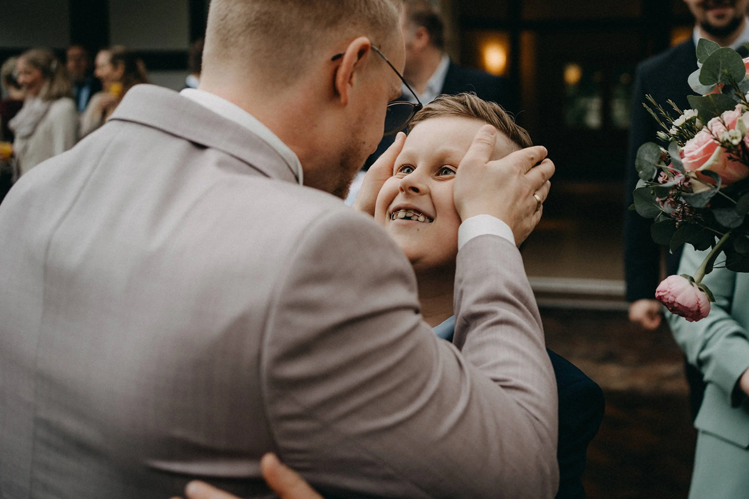 Ein Mann mit Brille und Kurzhaarfrisur hält die Hände eines lachenden Jungen, der im Mittelpunkt steht, in einem festlichen Raum. Im Hintergrund sieht man andere Personen, die feiern, und es gibt einen großen Blumenstrauß.