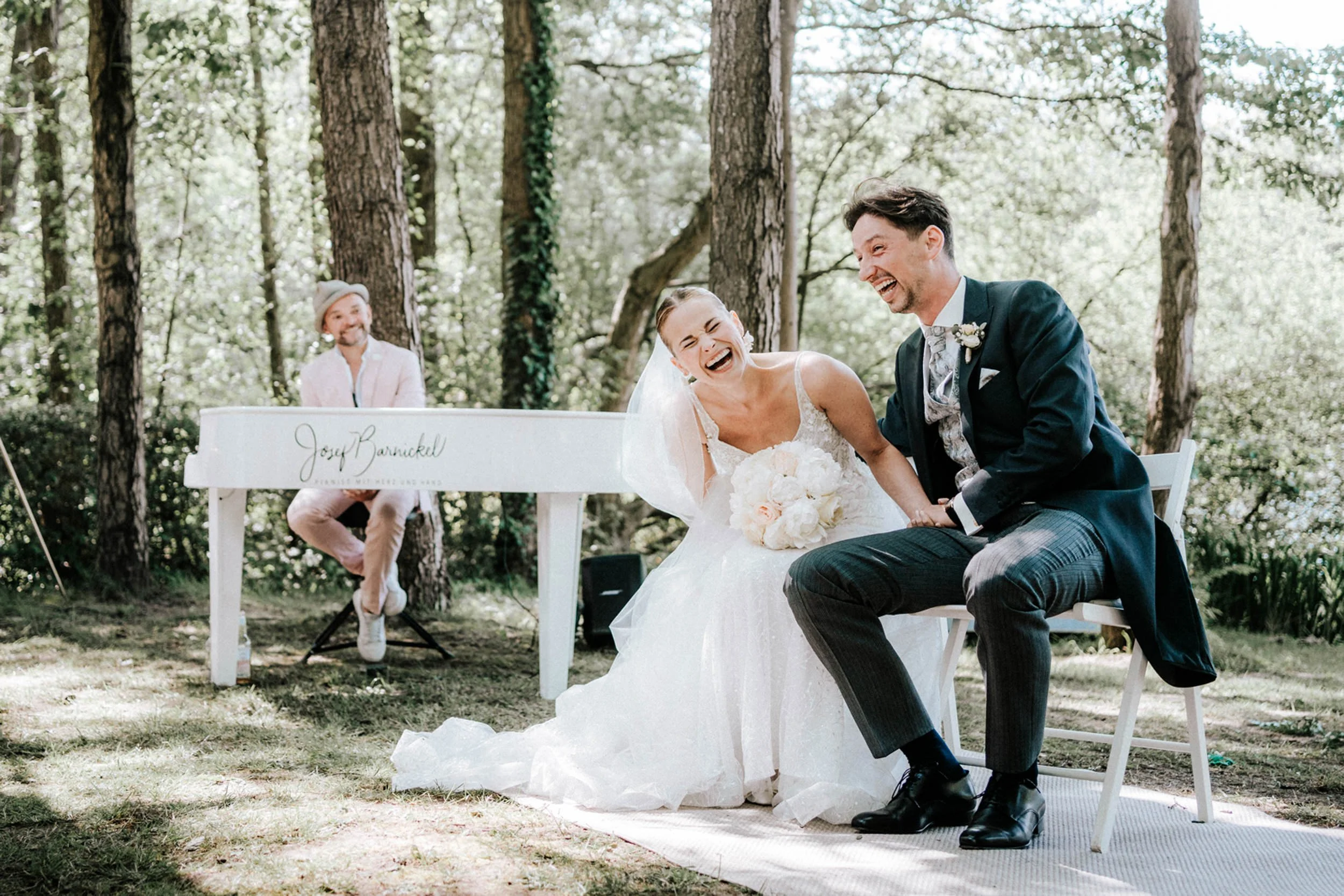 Ein lachendes Brautpaar bei einer Hochzeit im Freien, umgeben von Bäumen, mit einem Mann im Hintergrund, der auf einer weißen Bank sitzt.