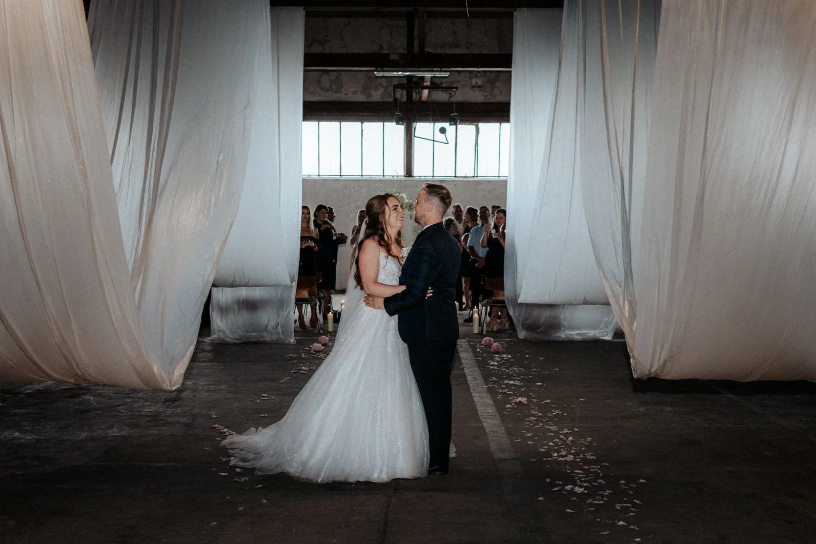 Ein Brautpaar tanzt bei ihrer Hochzeit, umgeben von Gästen und Dekorationen in einem-industrieähnlichen Raum, mit weißen Vorhängen und Kerzen auf dem Boden.