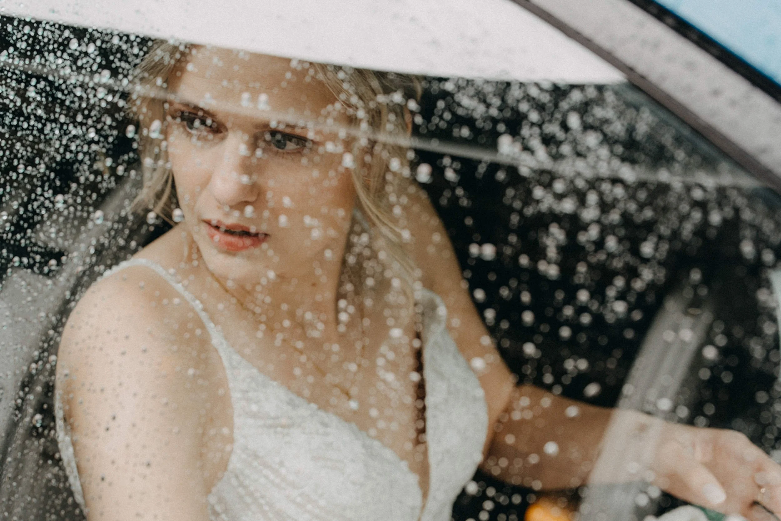 Eine blonde Frau in einem weißen Kleid wird hinter einer regennassen Autoscheibe beobachtet, wobei Regentropfen die Sicht verzerren.