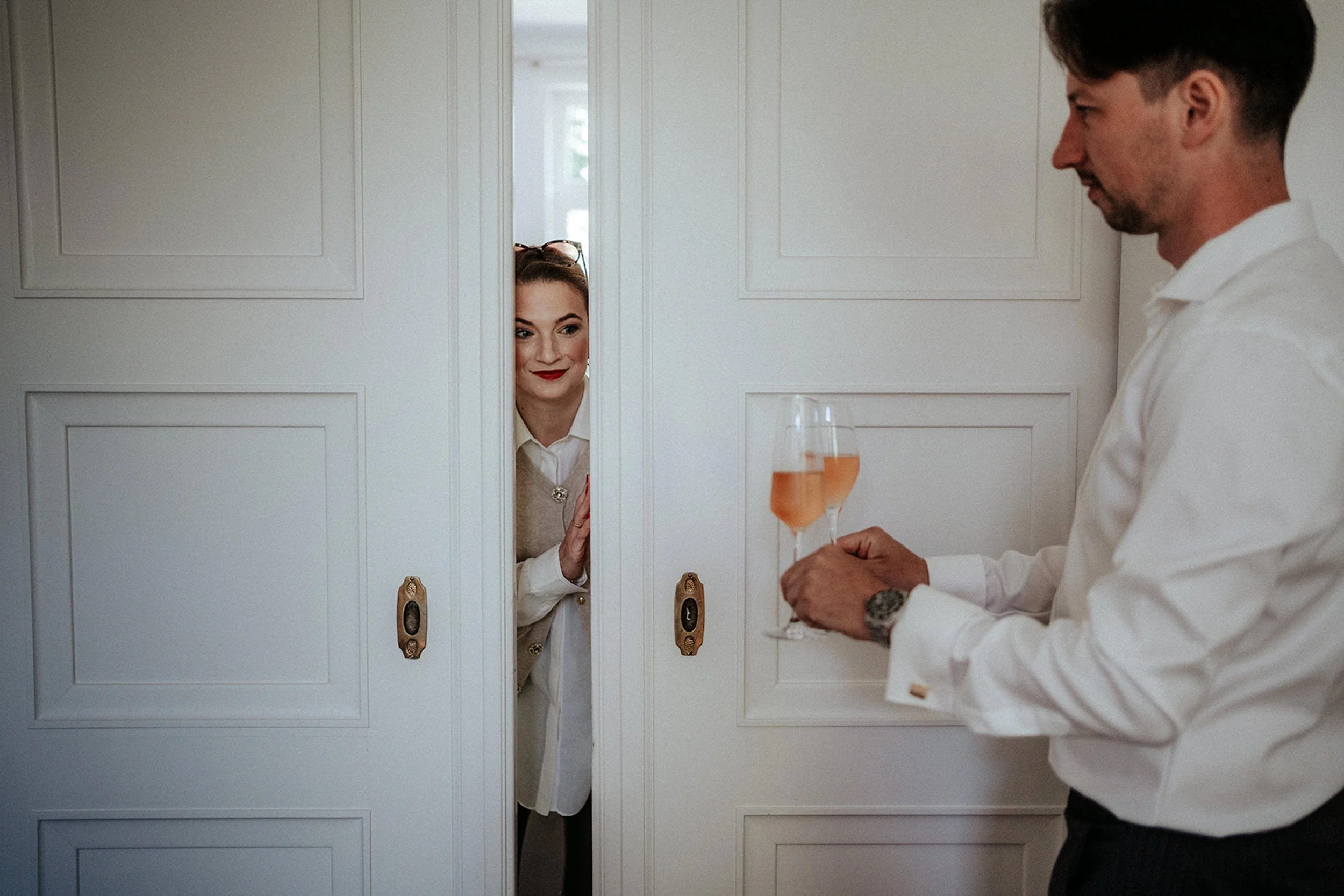 Ein Mann hält ein Glas Roséwein und steht vor einem weißen Schrank. Im Schrank schaut eine Frau mit roten Lippen und Brille durch eine kleine Öffnung in der Tür.
