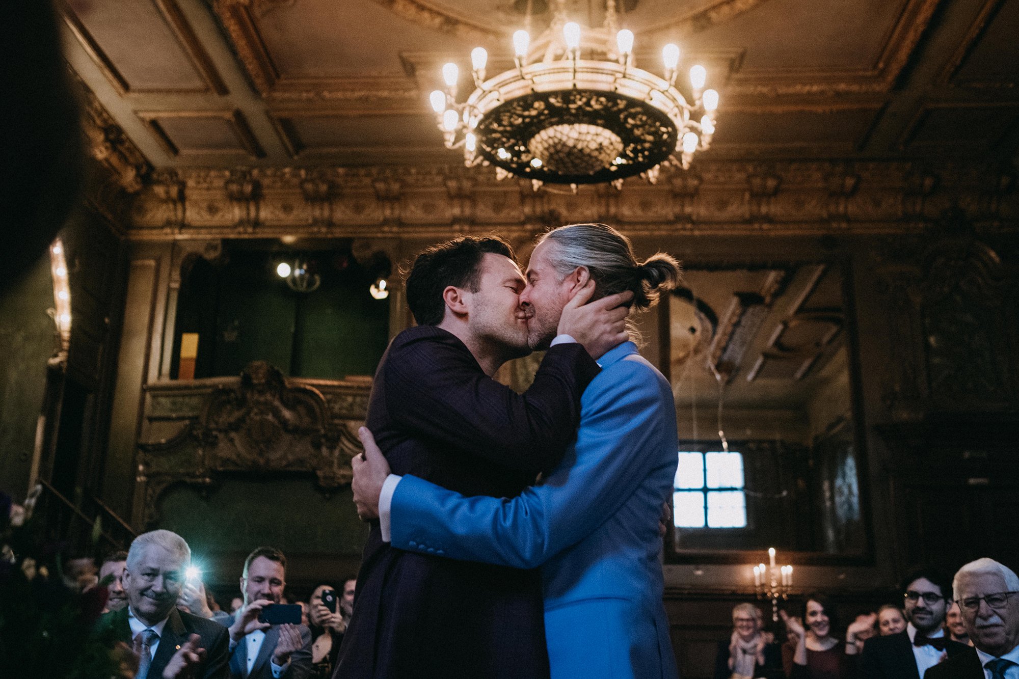 Zwei Männer tanzen eng umarmt in einem festlichen Raum. Die Szene wird von einem großen Kronleuchter beleuchtet. Im Hintergrund sieht man eine Gruppe von Menschen, die den Moment mit Fotos und Applaus feiern.