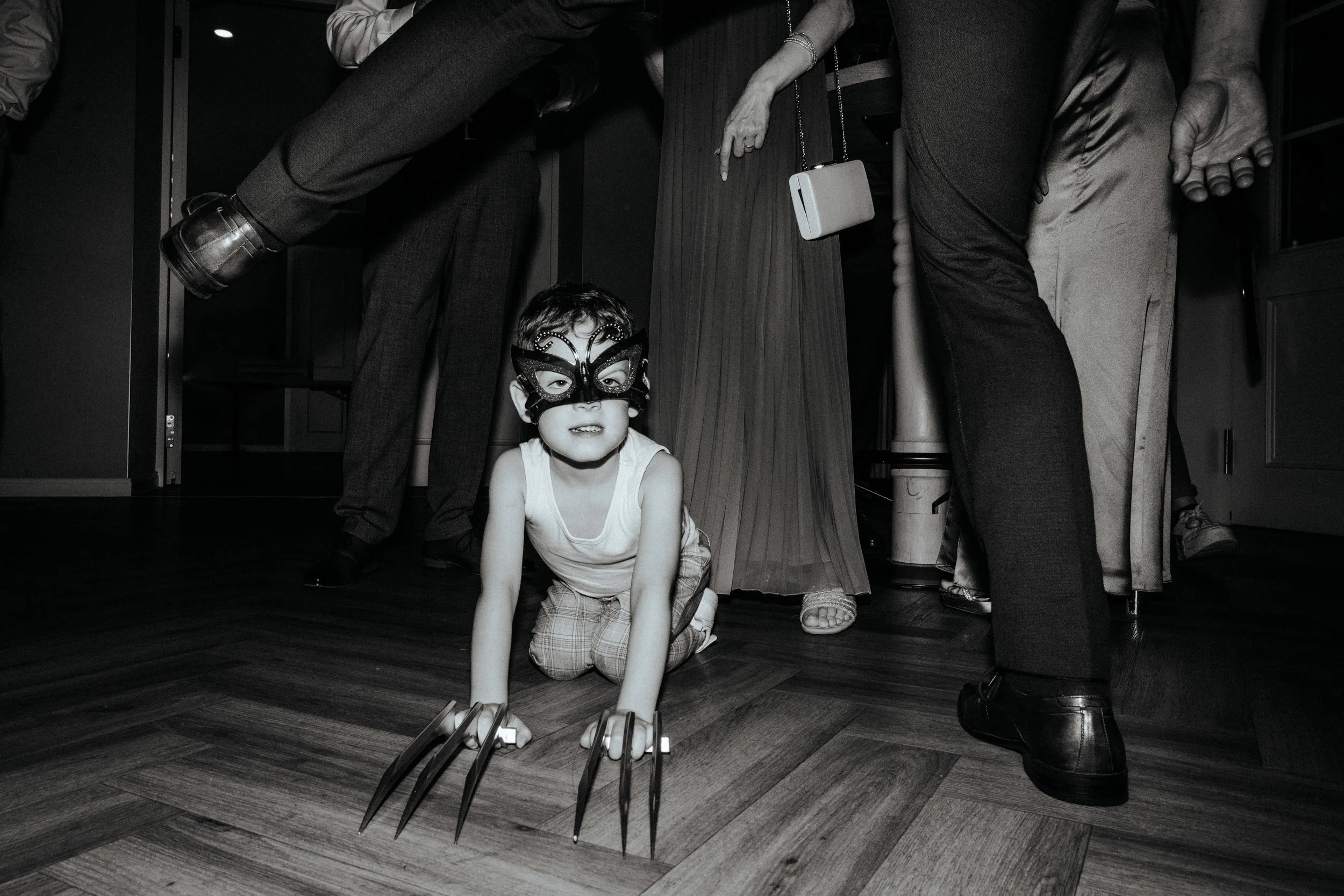 Ein Kind trägt eine Katzenmaske und ist auf den Knien, während Erwachsene um ihn herumstehen, einige mit ausgestreckten Beinen und Schuhen. Die Szene ist schwarz-weiß aufgenommen.