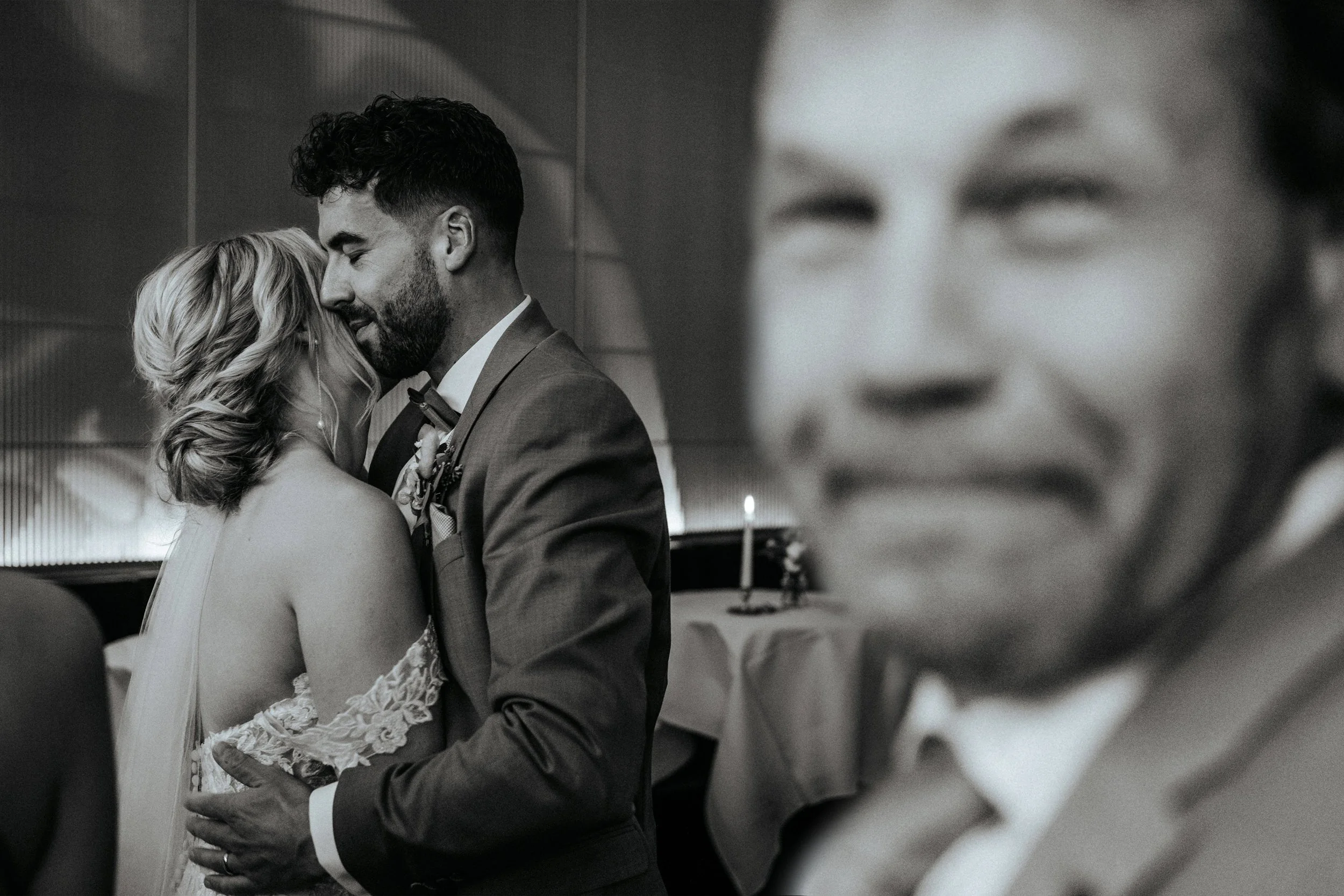 Ein Paar bei einer Umarmung, vermutlich auf einer Hochzeit, mit einem lachenden Mann in einem Anzug und einer Frau in einem weißen Hochzeitskleid mit Spitze, während im Hintergrund eine große Leinwand mit einem lachenden Gesicht zu sehen ist.