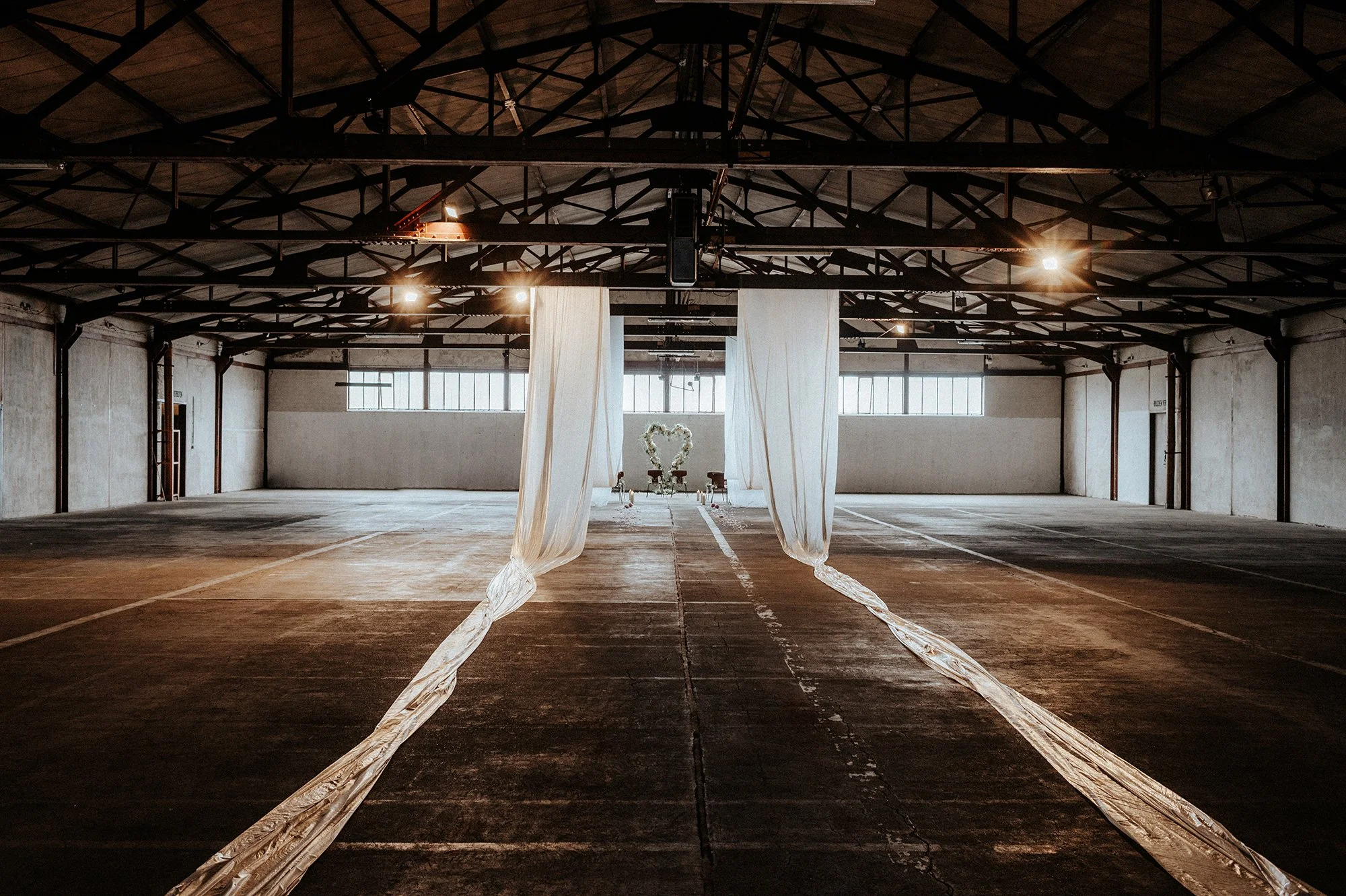 Ein leerer Industriehalle mit hohen Decken und großen Fenstern. Zwei weiße Stoffvorhänge hängen an der Mitte, in der Nähe befindet sich eine romantische Herzdekoration, vermutlich für eine Hochzeit.