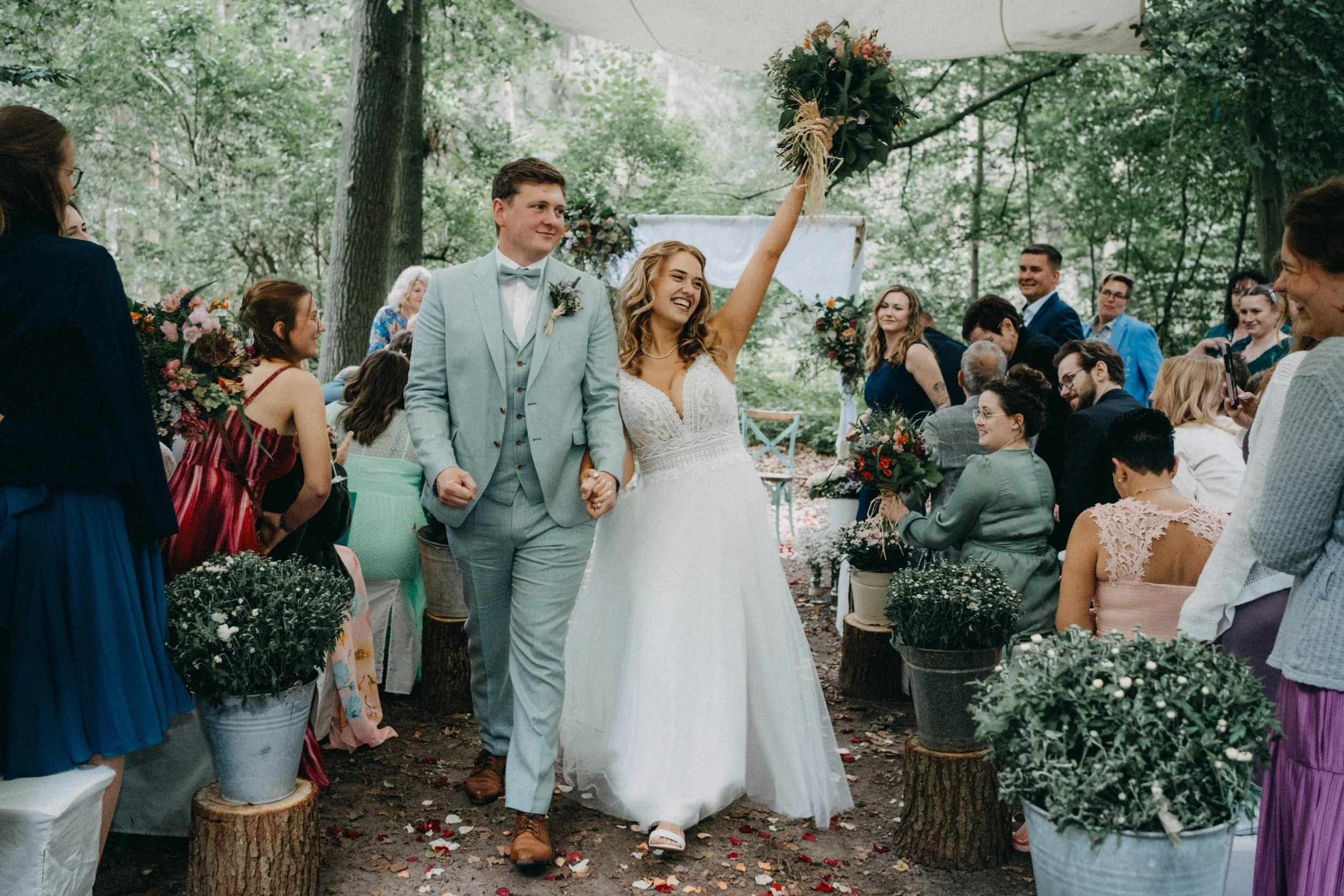 Ein glückliches Brautpaar bei einer Hochzeit im Freien, umgeben von Gästen, mit Blumen und Bäumen in der Natur.