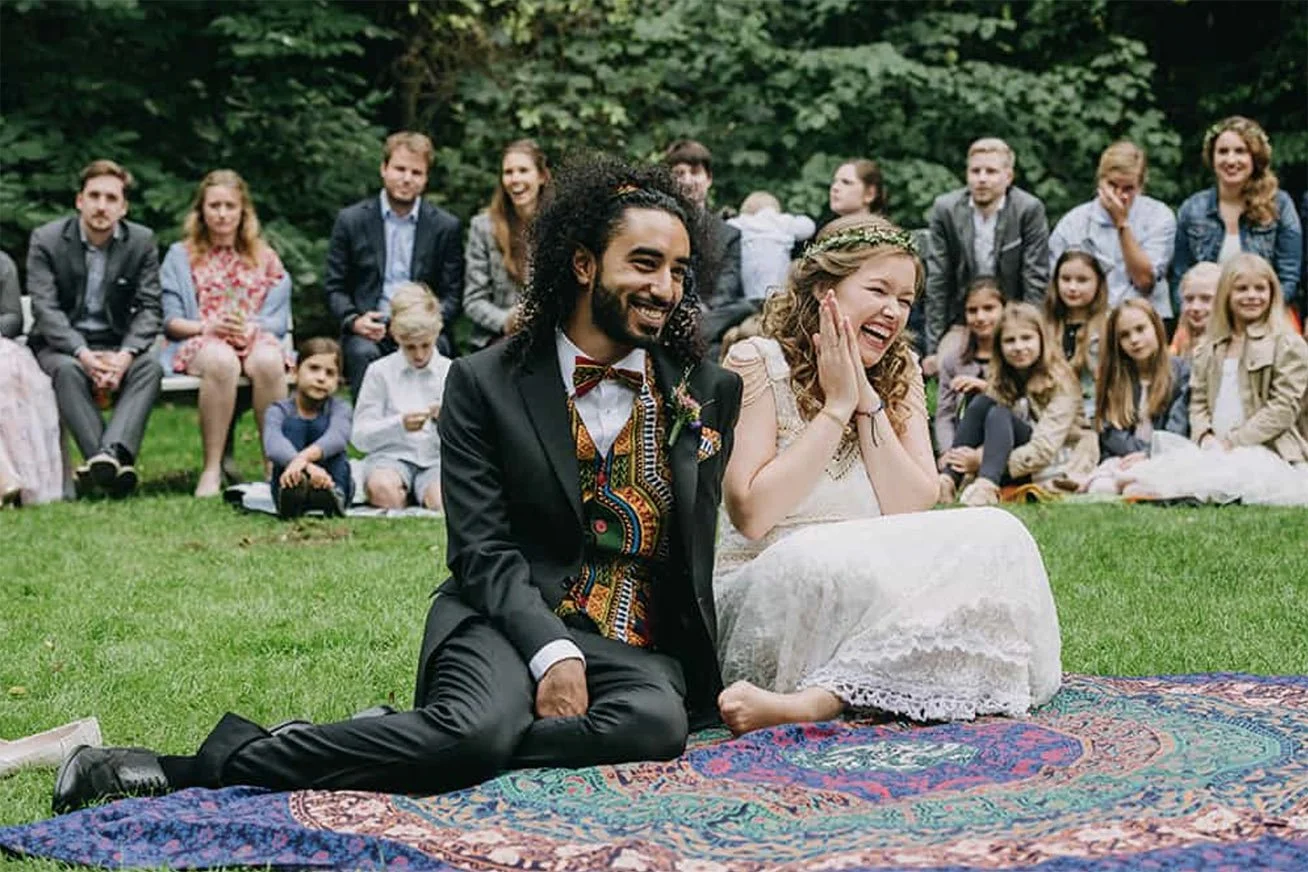 Ein Paar sitzt lachend auf einem bunten Teppich im Freien bei einer Hochzeit, umgeben von lachenden Gästen.