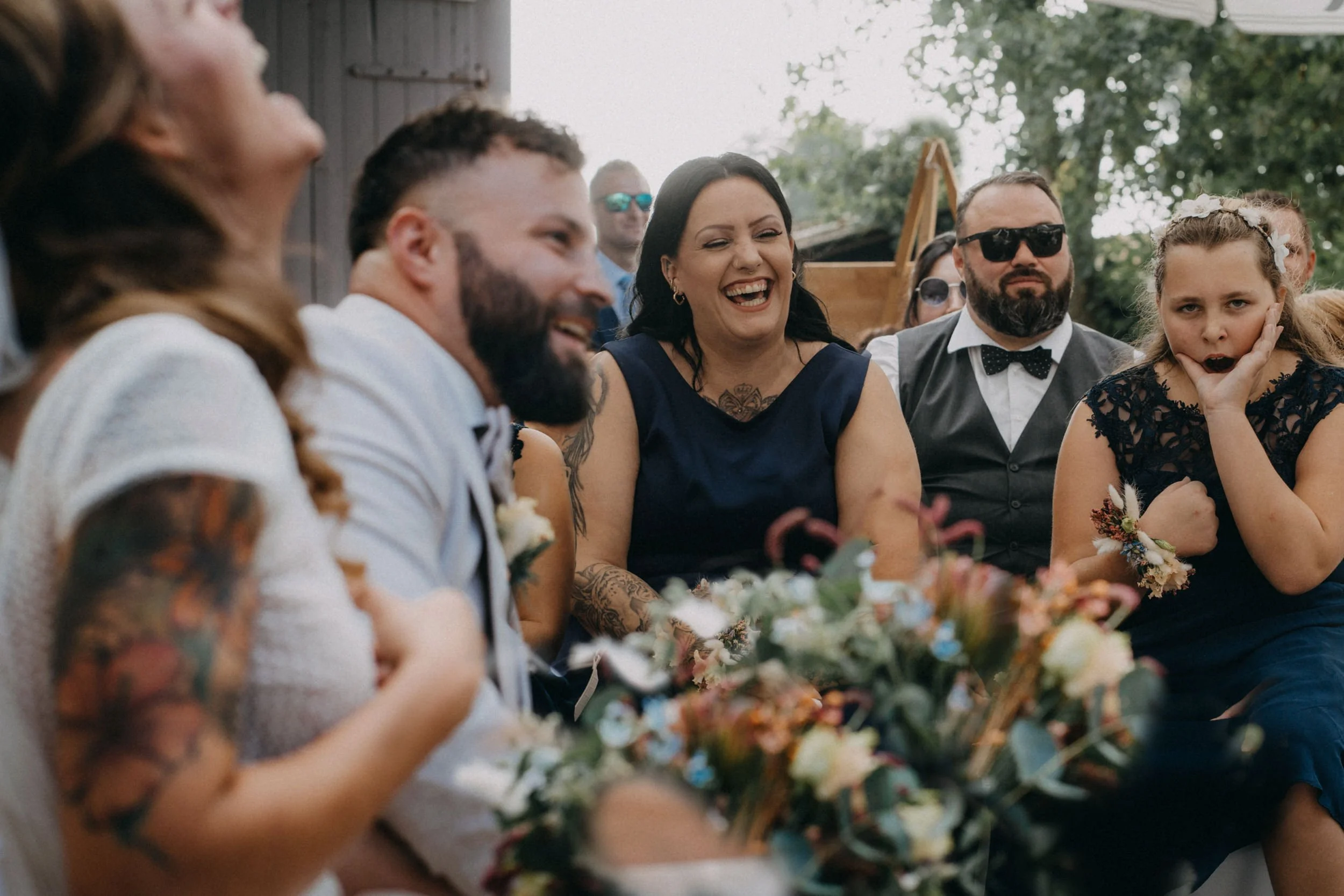 Gruppe von Personen bei einer Hochzeit, sie lachen und unterhalten sich im Freien mit Bäumen im Hintergrund.