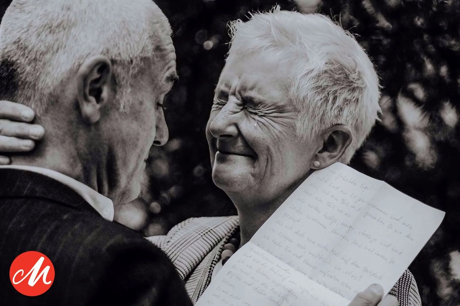 Ein älteres Paar umarmt sich liebevoll, die Frau hält ein handgeschriebenes Blatt Papier. Beide lächeln emotional.