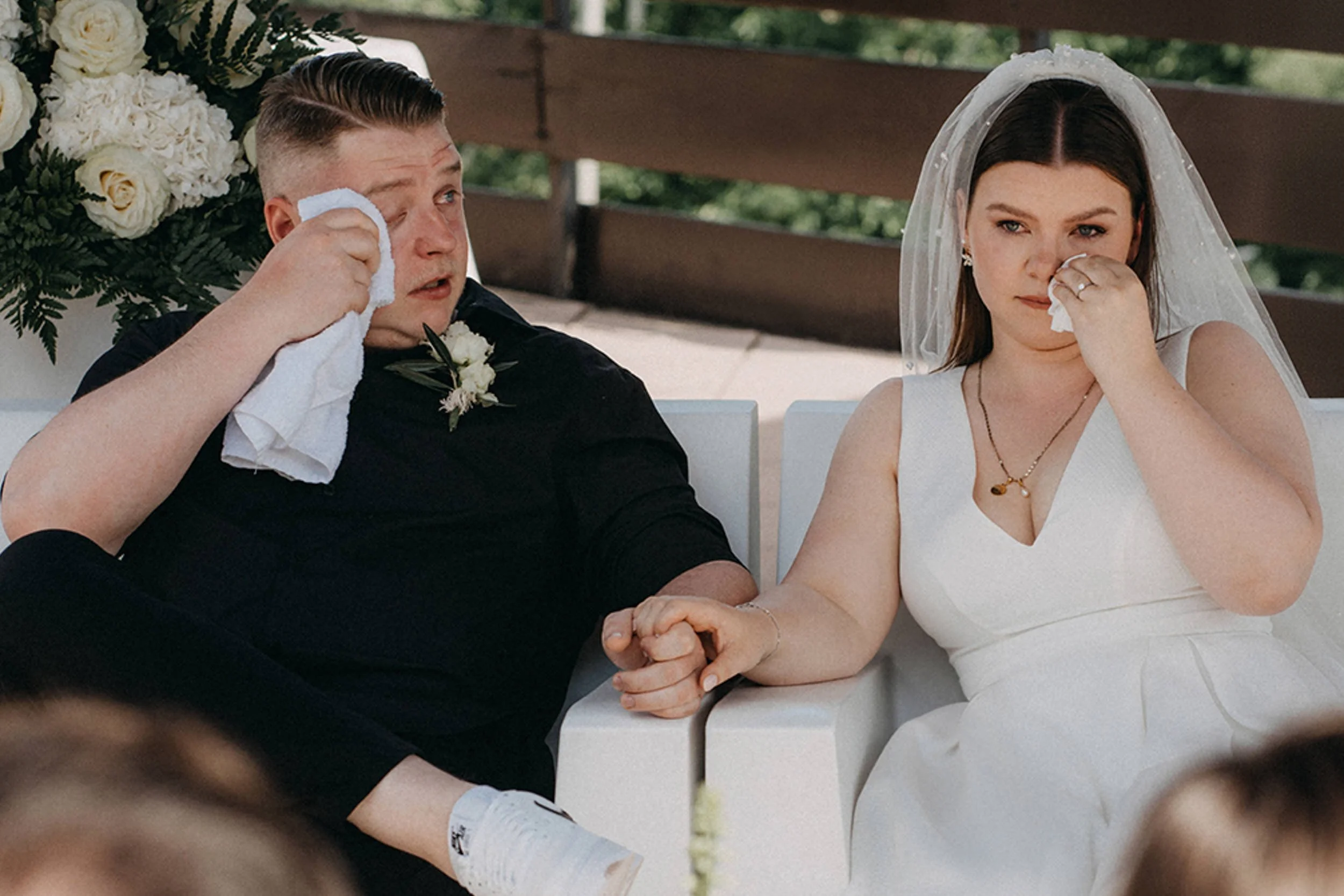 Ein Brautpaar sitzt bei ihrer Hochzeit, beide weinen und halten Händchen, die Braut trägt ein weißes Kleid und einen Schleier, der Bräutigam trägt einen schwarzen Anzug, im Hintergrund sind weiße Blumen und grüne Blätter zu sehen.