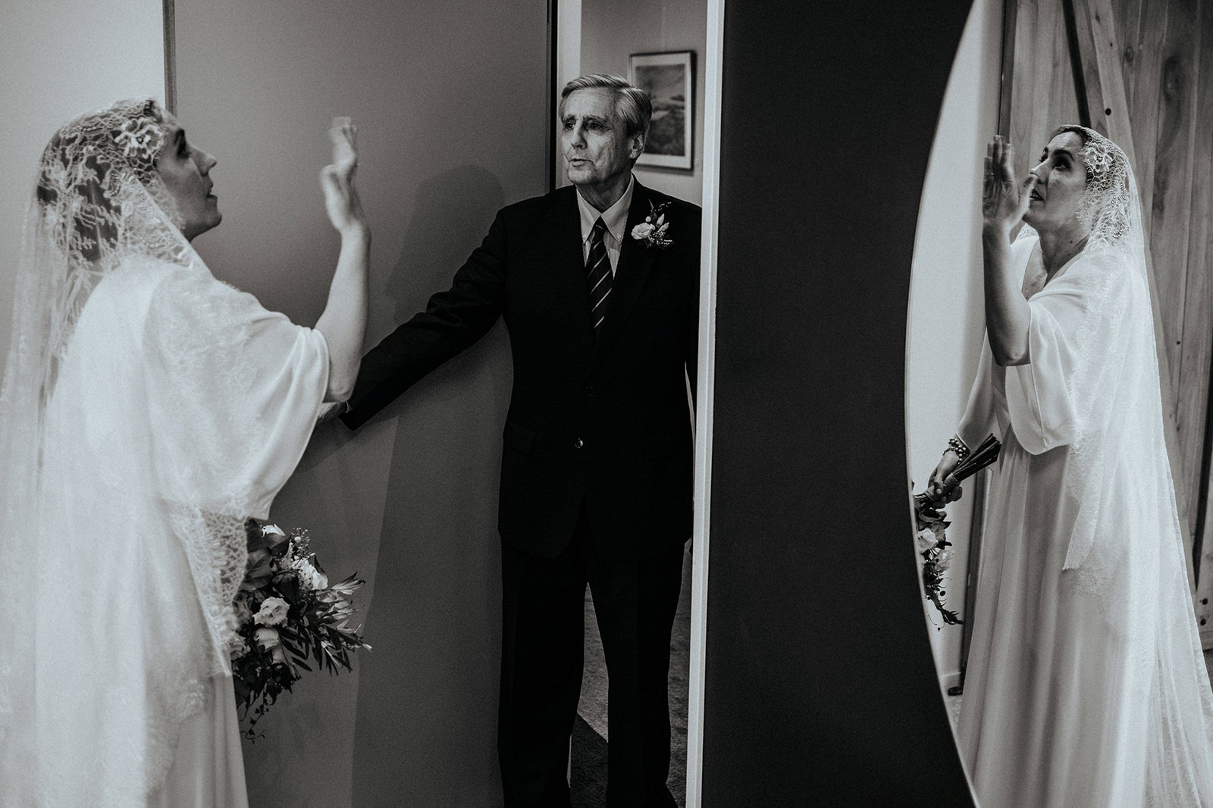 Zwei Frauen im Brautkleid stehen sich gegenüber und berühren sich an den Händen, während ein älterer Mann zwischen ihnen steht, vermutlich bei einer Hochzeitszeremonie.
