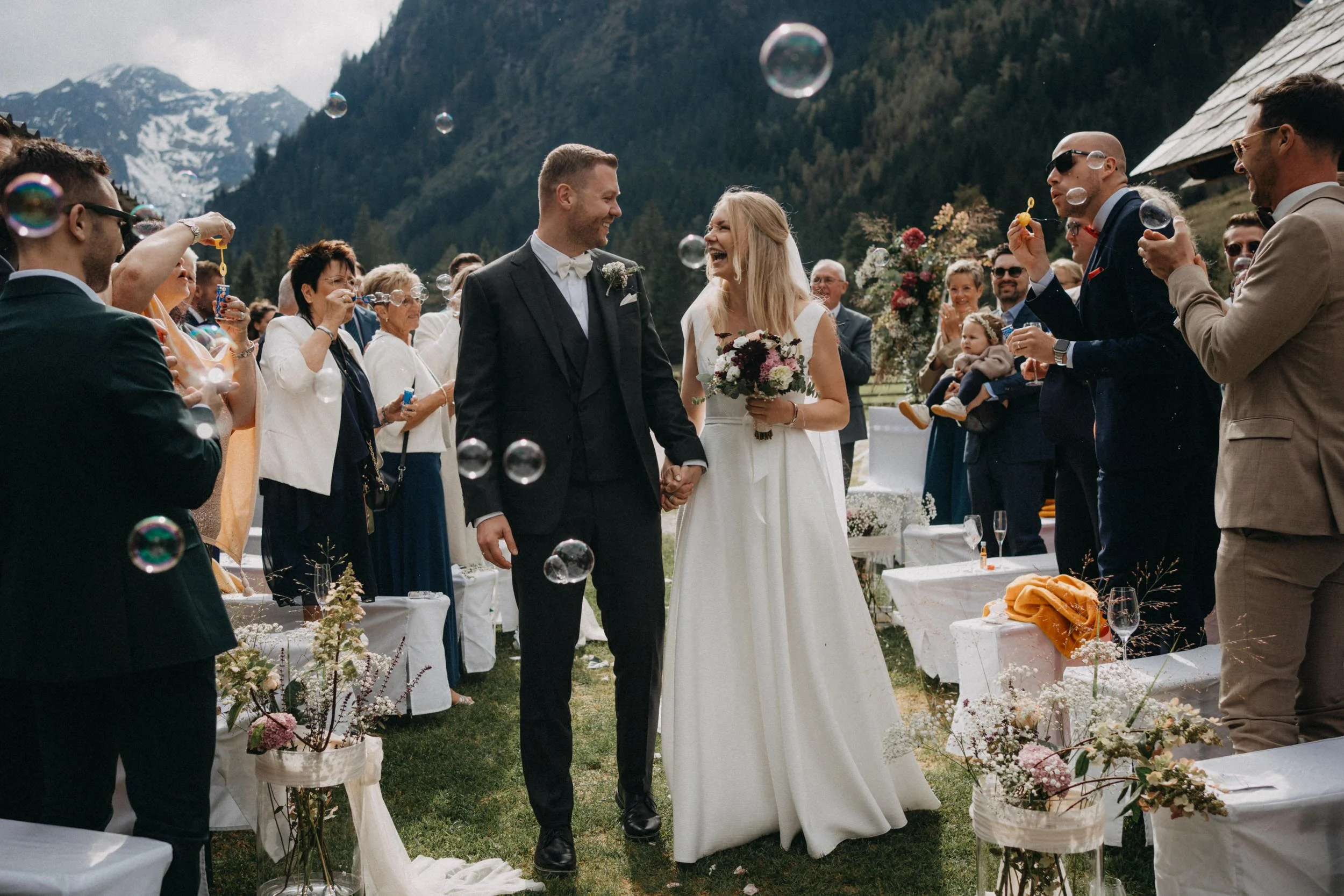 Ein Brautpaar bei einer Hochzeit im Freien mit Bergen im Hintergrund, umgeben von Gästen, die Seifenblasen machen.