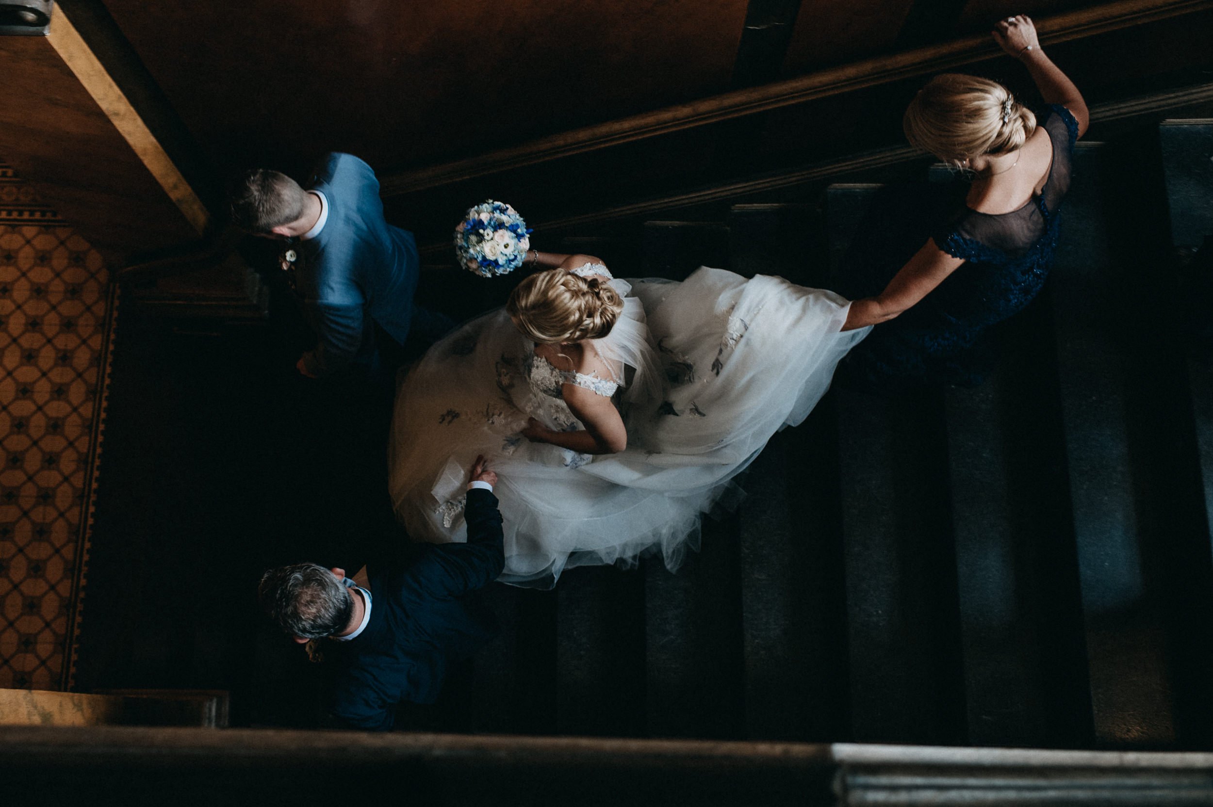 Hochzeitspaar, sichtbar von oben, auf einer Treppe, umgeben von mehreren Personen, die vermutlich Gäste sind, in einem eleganten Innendesign.