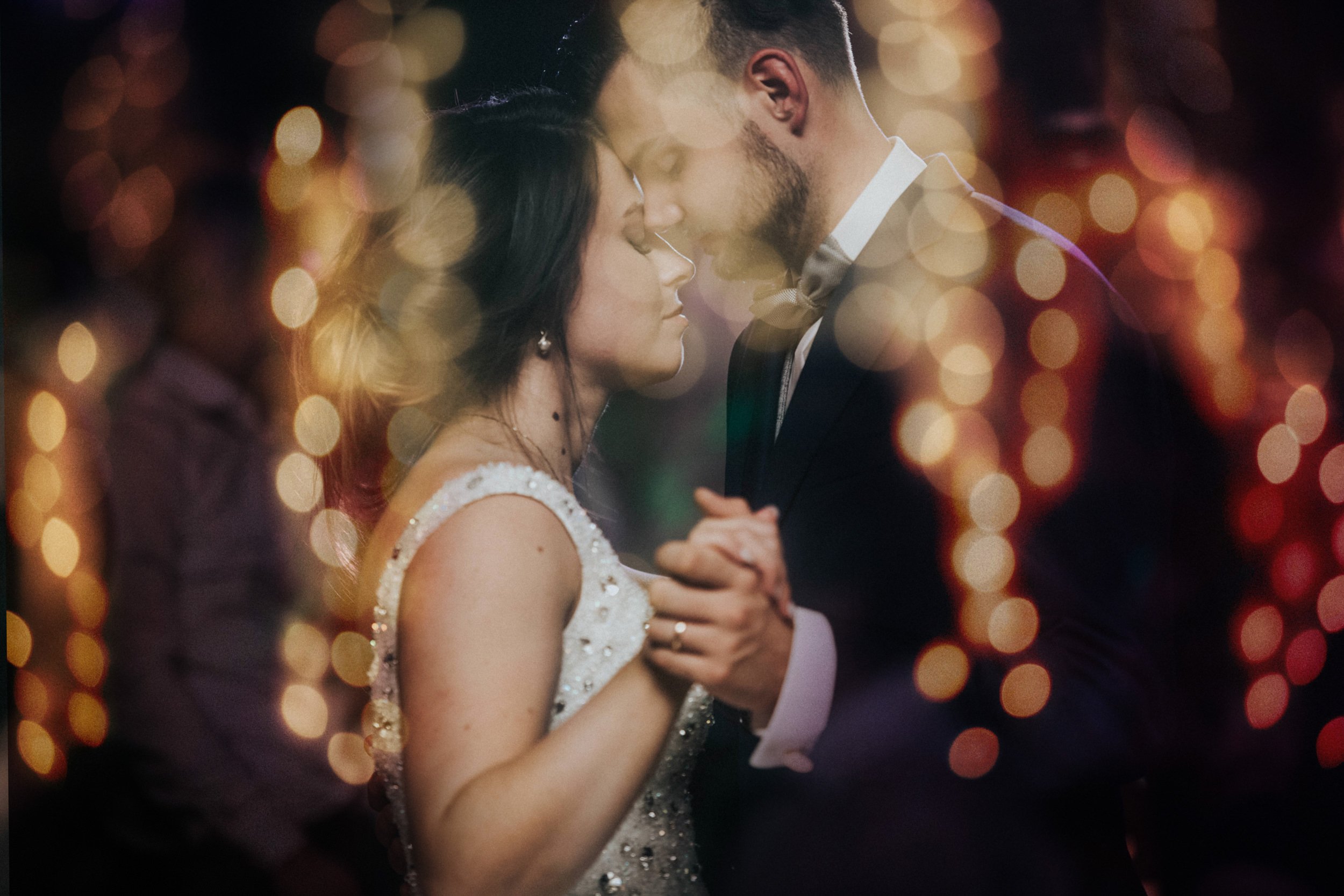 Ein Paar tanzt auf einer Hochzeit bei Kerzenlicht, im Hintergrund sind bunte Lichter und verschwommene Gäste.