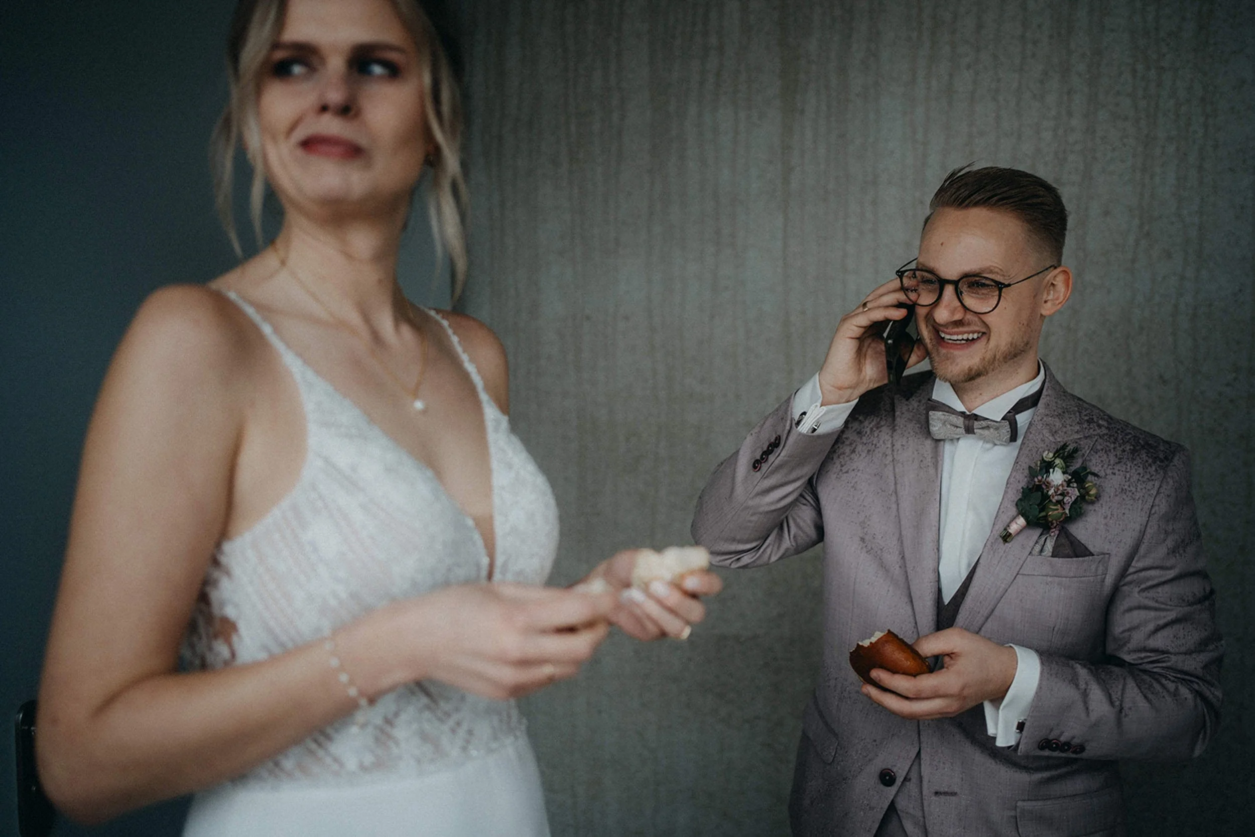 Ein Mann und eine Frau bei einer Hochzeit, der Mann trägt einen grauen Anzug mit Fliege und die Frau trägt ein weißes Brautkleid, während sie etwas in den Händen hält. Der Mann spricht am Telefon, die Frau schaut verlegen.