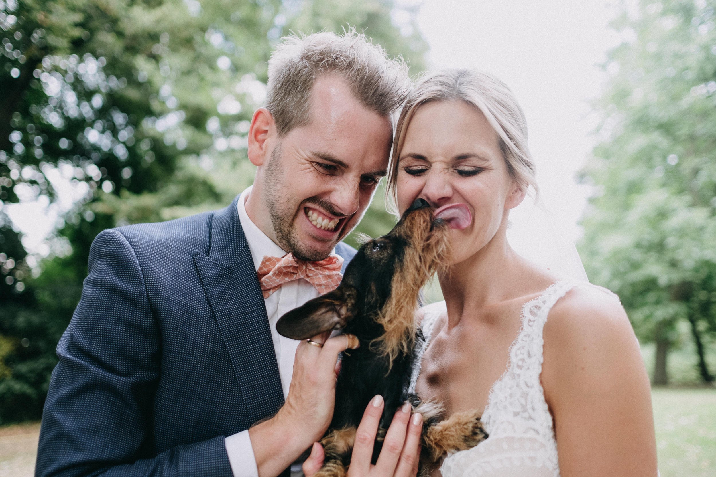 Ein Mann und eine Frau bei einer Hochzeit, die einen kleinen Hund streicheln und küssen, umgeben von grünen Bäumen im Freien.