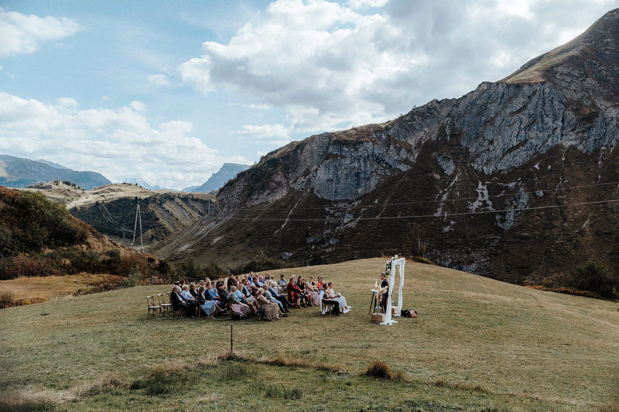 Eine Gruppe von Menschen sitzt bei einer Outdoor-Hochzeit vor einem Bergblick, die Landschaft ist mountainous mit Wolken am Himmel.