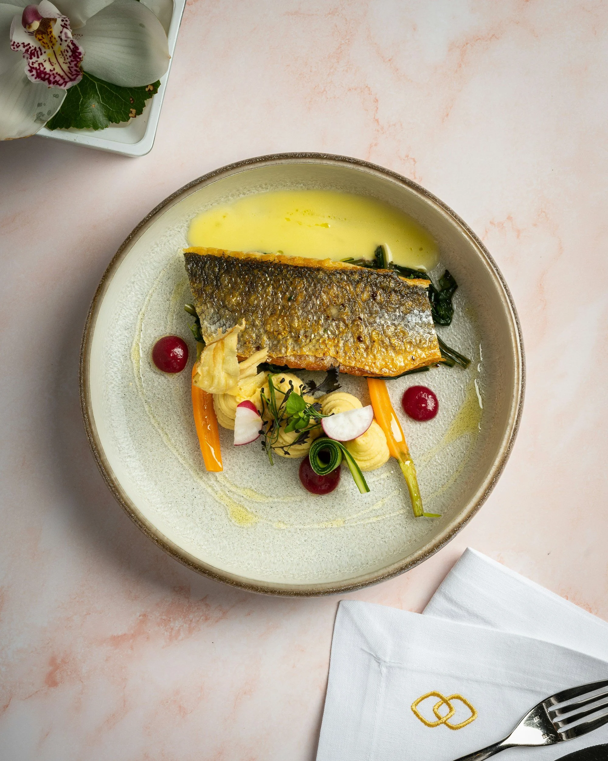 A plated dish featuring a piece of cooked fish with crispy skin, served with a yellow sauce, garnished with vegetables, herbs, and small red berries, on a round beige plate on a pink table.