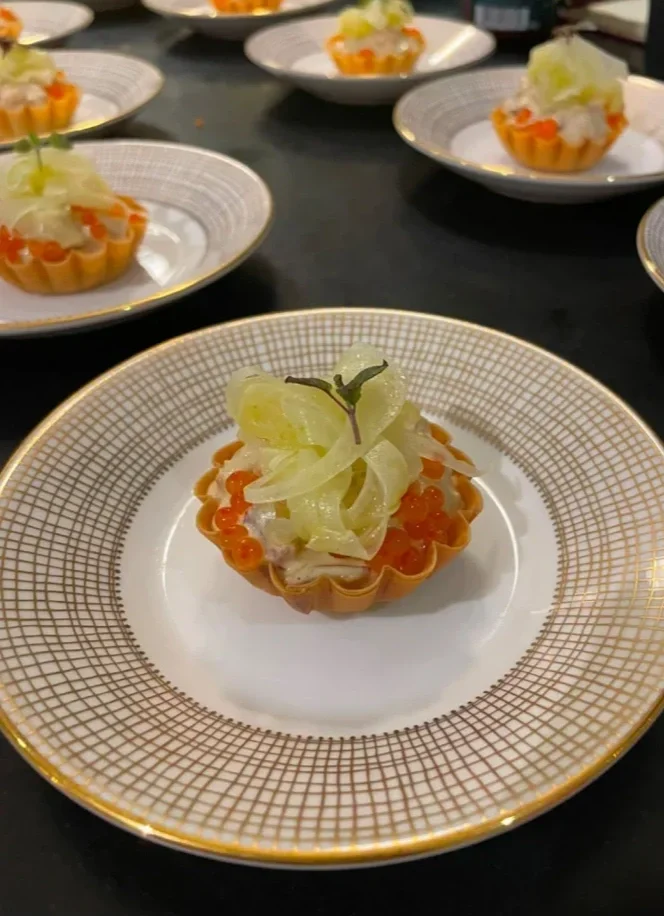 A small tart shell topped with white seafood, orange fish roe, and garnished with thinly sliced fennel and a tiny herb leaf, served on a decorative porcelain plate.