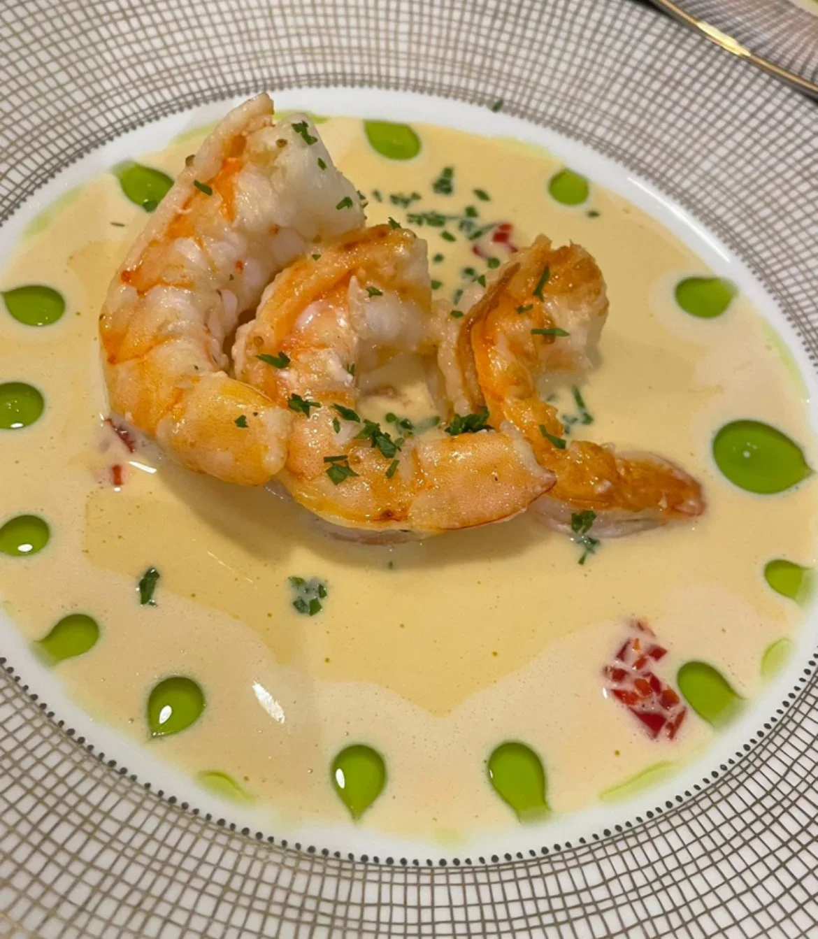 Shrimp in a creamy sauce garnished with chopped herbs and surrounded by green oil drops on a patterned plate.