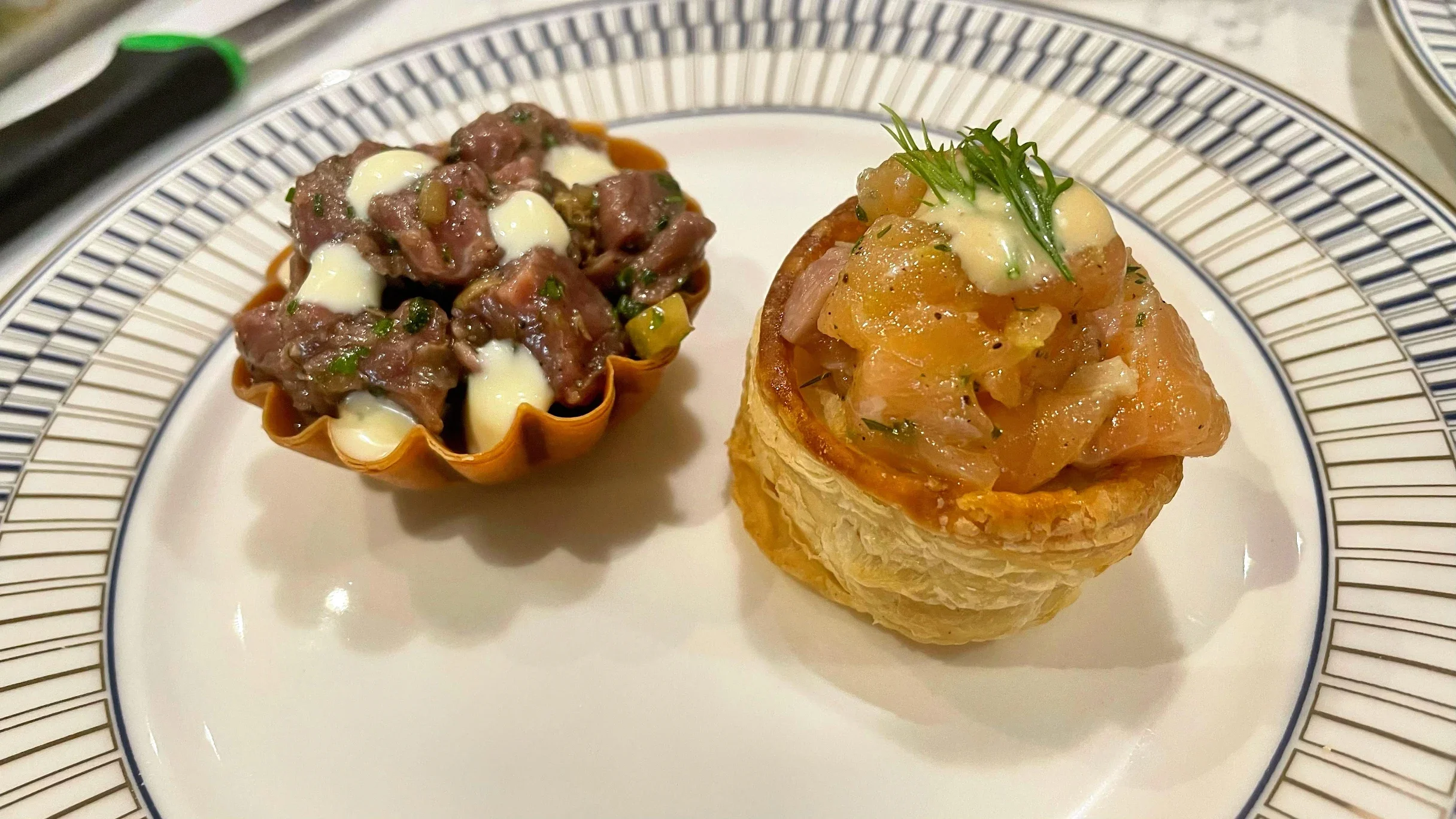 Two gourmet appetizers on a decorative plate: one is a tart shell stuffed with steak tartare topped with cream, the other is a puff pastry cup filled with ceviche topped with cream and herbs.