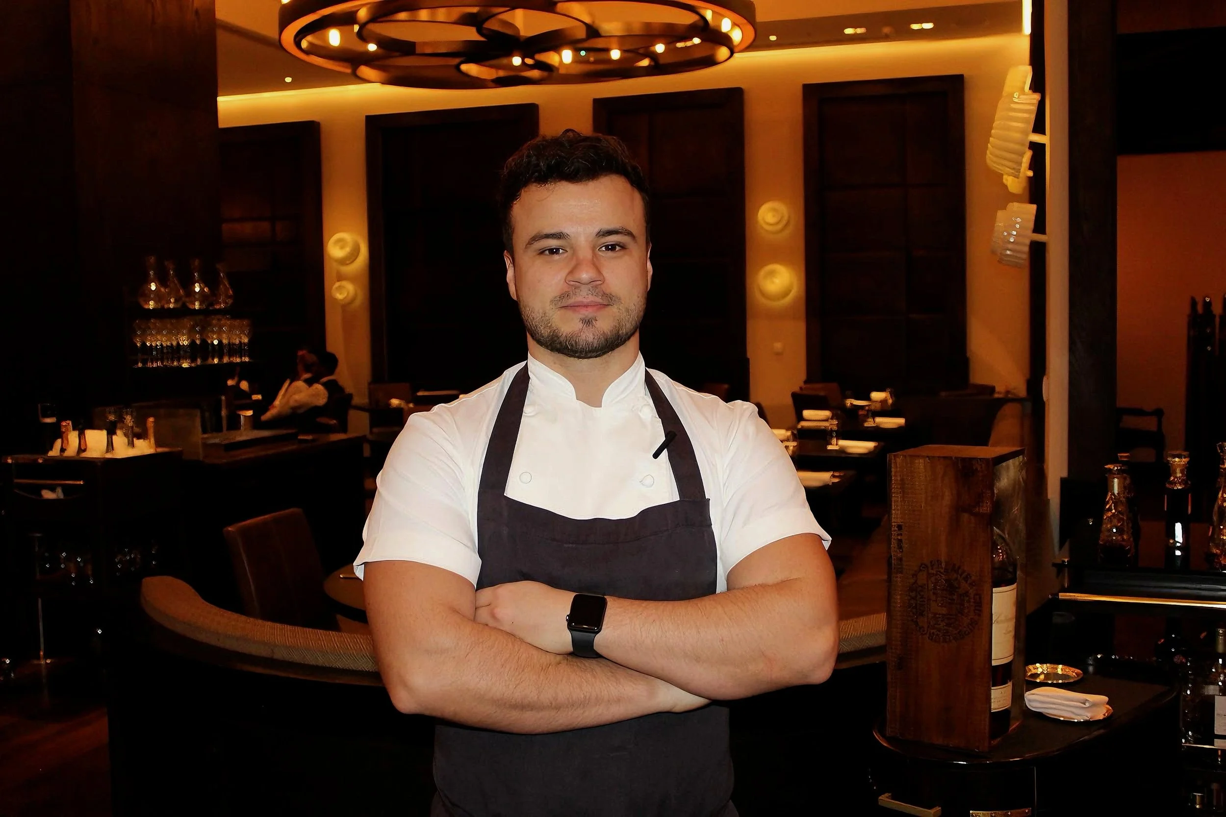 A male chef standing with arms crossed in a restaurant, wearing a white chef's coat and a dark apron, with a smartwatch on his left wrist. The restaurant has dim lighting, tables set with cutlery, and modern decor.