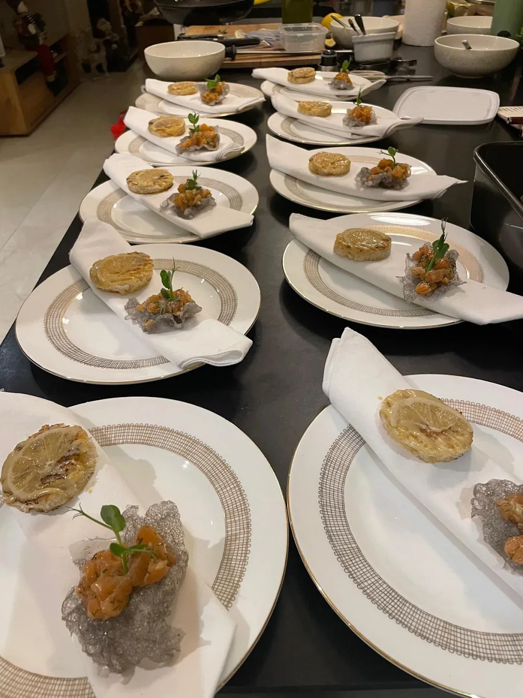 Several white porcelain plates with ornate gold trim, each holding a folded white napkin filled with a small round pastry topped with lemon slices and a small green garnish, and a portion of a crispy gray rice cracker with an orange topping and a gre