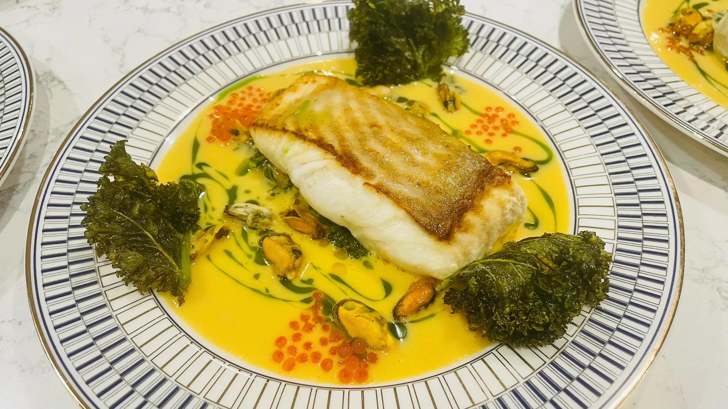 A plated dish of seared white fish fillet on a bed of vibrant yellow sauce with green and red accents, garnished with greens and roe, served on a decorative white and black patterned plate.