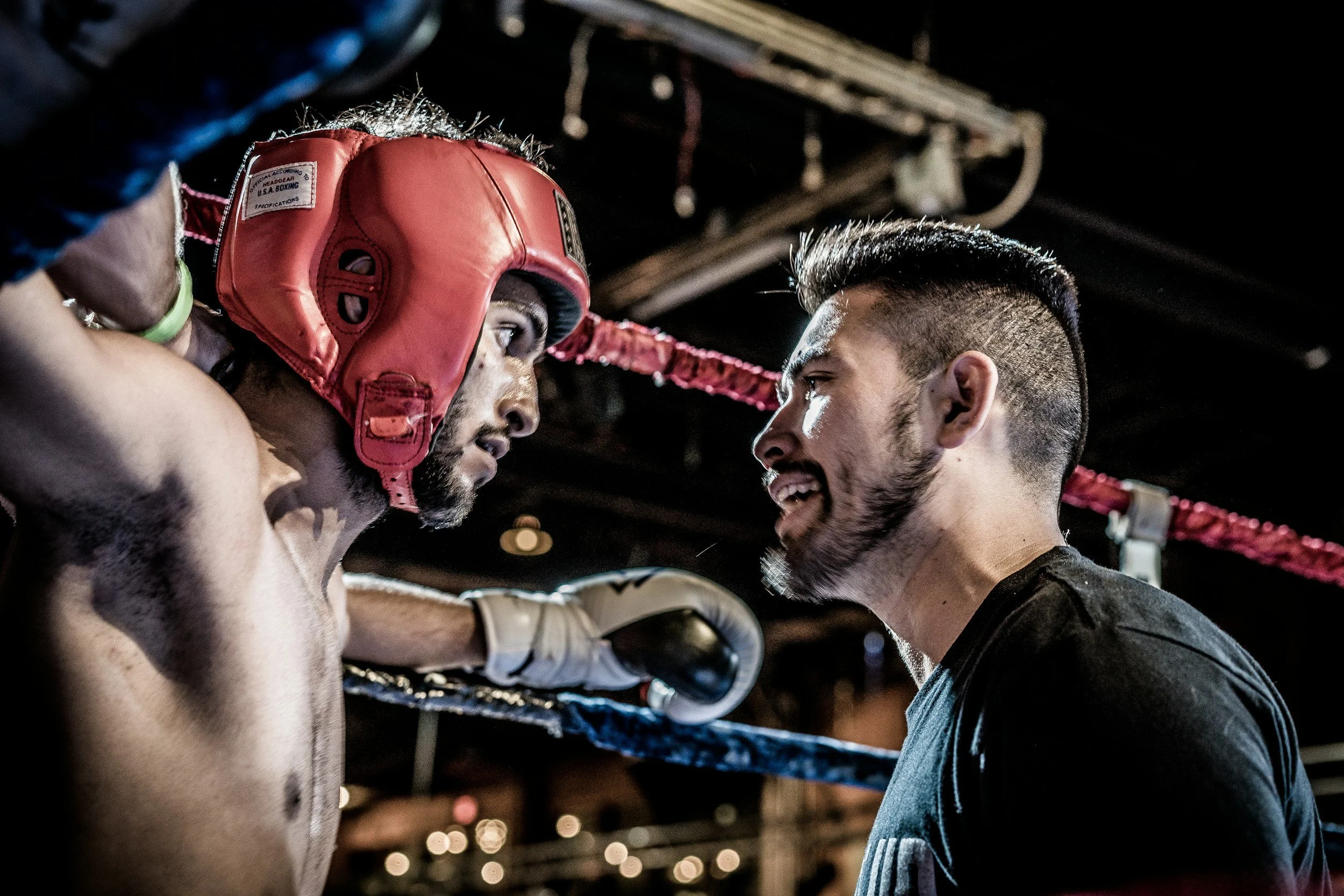 A boxer with a red headgear facing a trainer inside a boxing ring, with the boxer appearing focused and the trainer speaking to him.