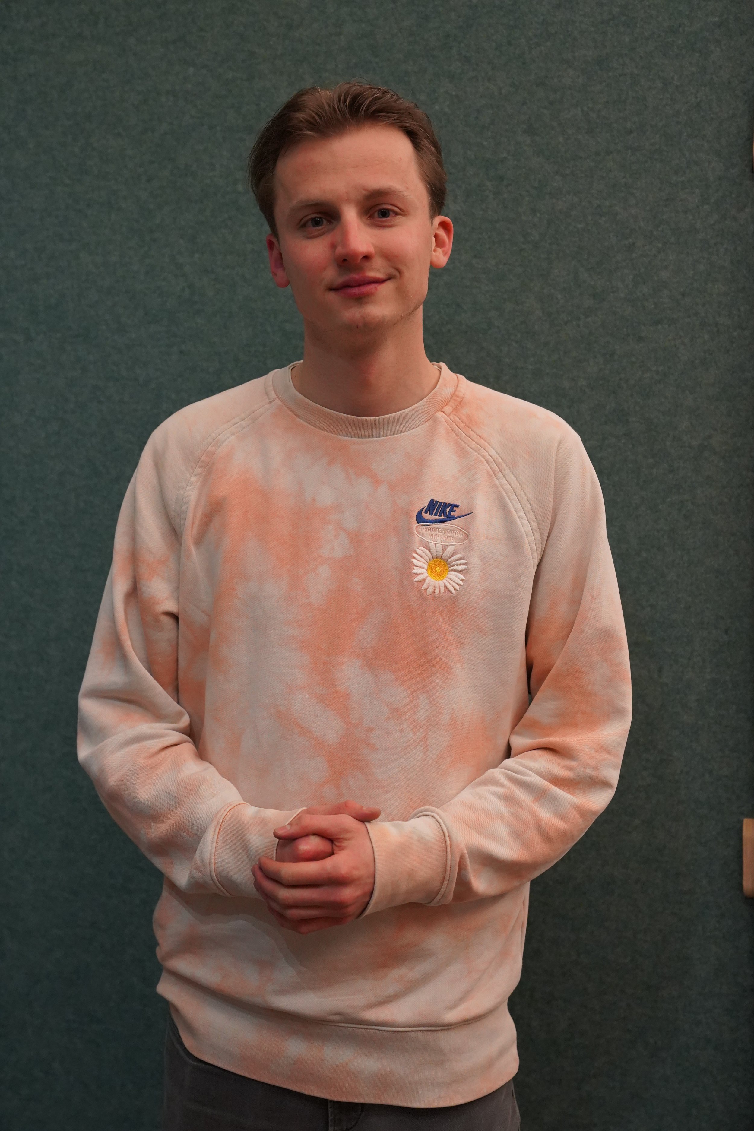 A young man with short brown hair and fair skin standing against a dark teal wall, wearing a light-colored, tie-dye sweatshirt with a Nike logo and a daisy flower embroidery on the chest. He is smiling slightly and has his hands clasped in front of him.
