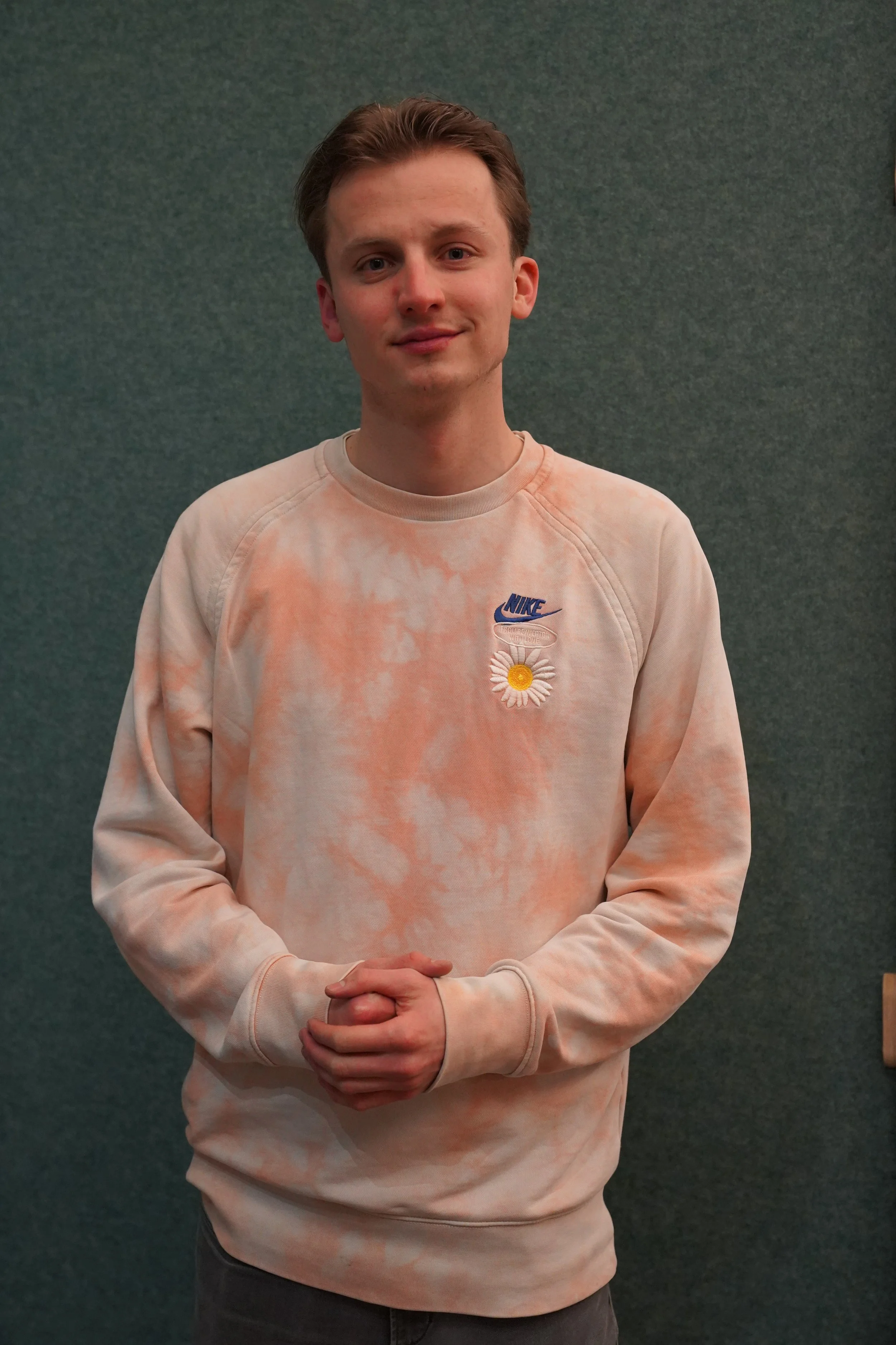 A young man with light skin and brown hair standing in front of a green textured wall, wearing a light orange and white tie-dye Nike sweatshirt with a daisy and coffee cup patch on the chest, smiling slightly with clasped hands.