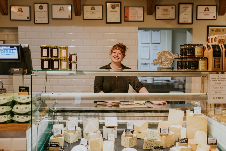 a woman standing behind a food counter filled with farmhouse cheese in a farm shop in scotland