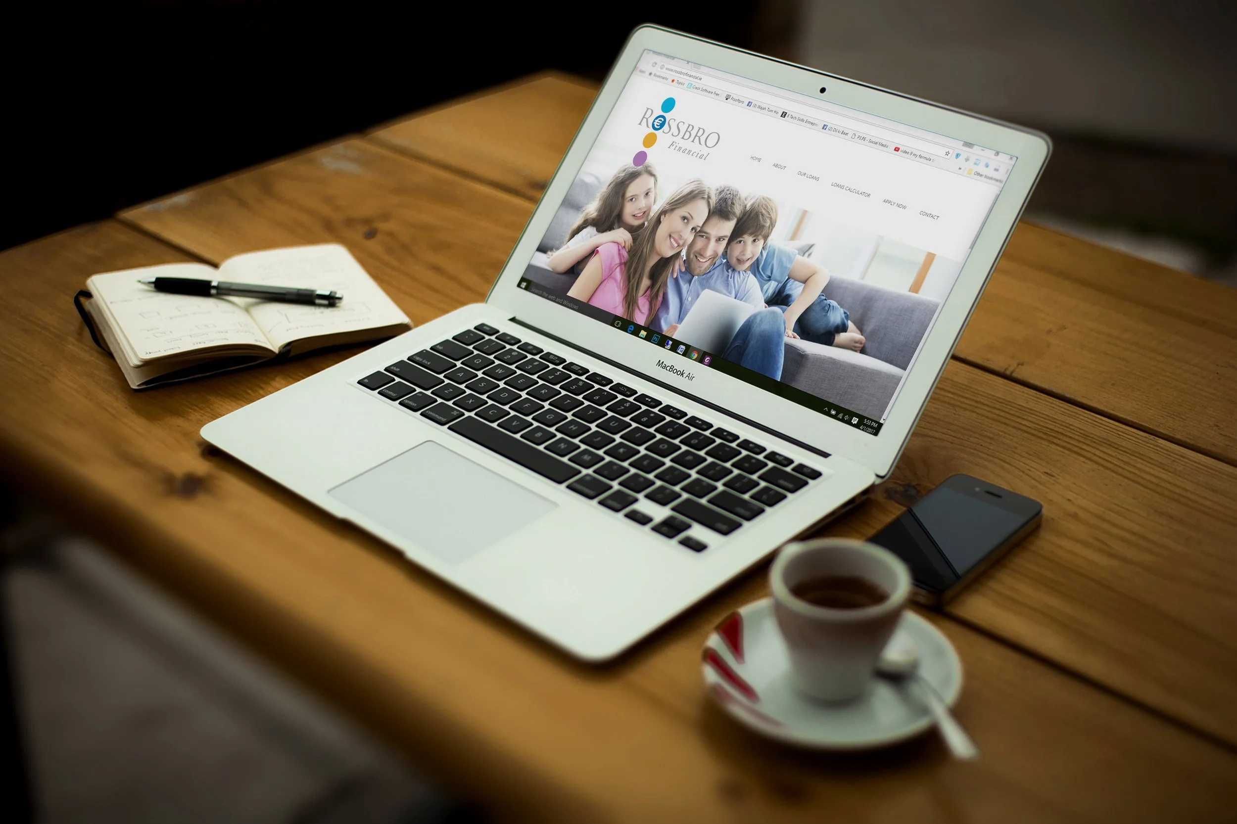 Open laptop on wooden table displaying a family on the Rossbro Financial website, with a notebook and pen, a smartphone, and a cup of coffee nearby. Display is to show where a customer would start the loan or lending process.